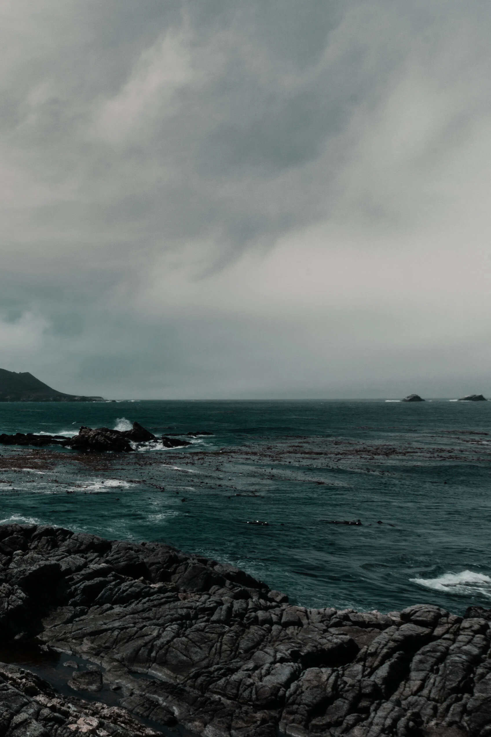 Dark teal sea crashes against black and grey rocks of California coast under cloudy sky.