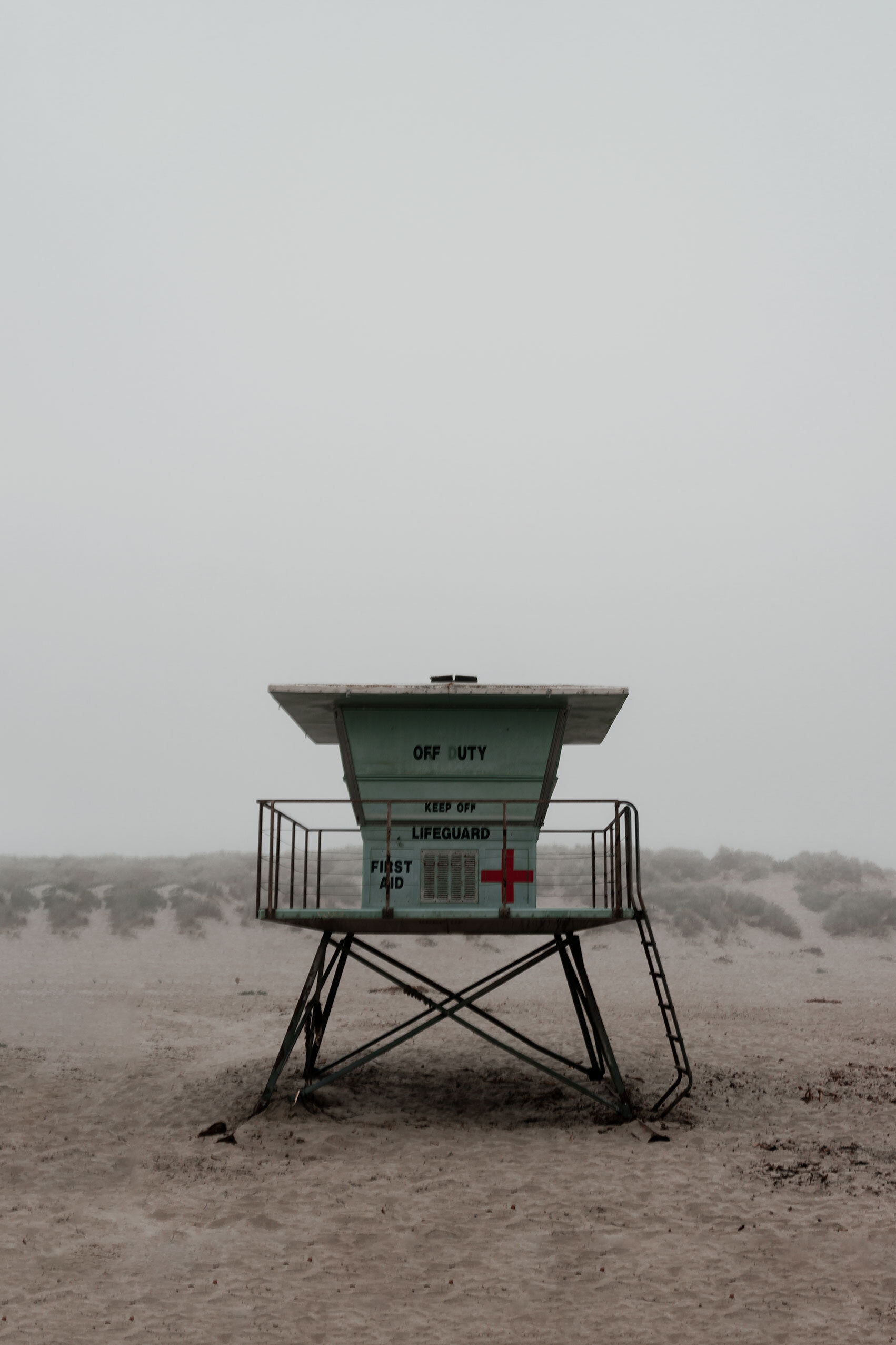 Light mint blue lifeguard building on stilts on a foggy California beach of light sand.