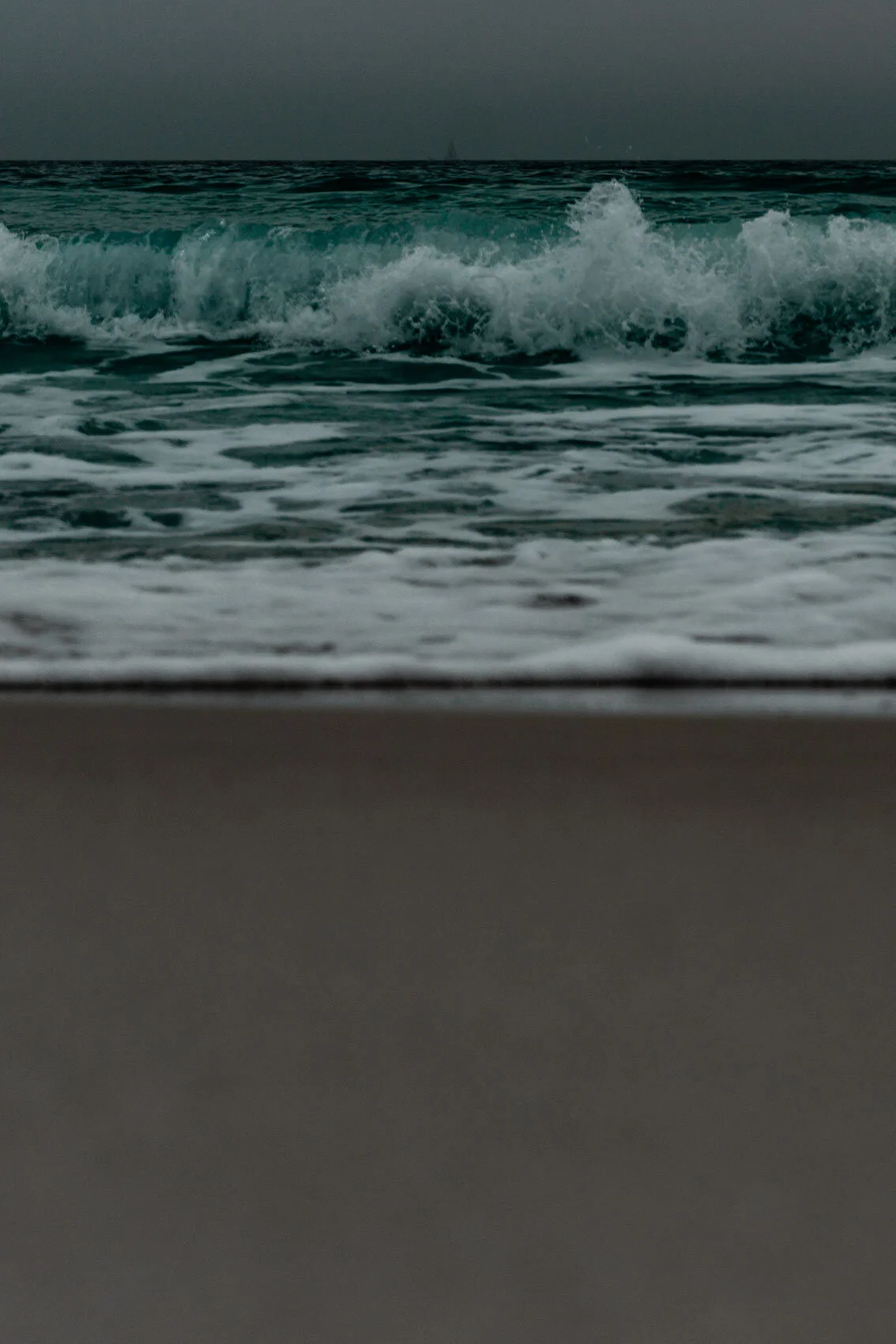 Bug's eye view of Florida beach. Grey sky, dark ocean, teal waves, white sea foam, and light sand form horizontal stripes.