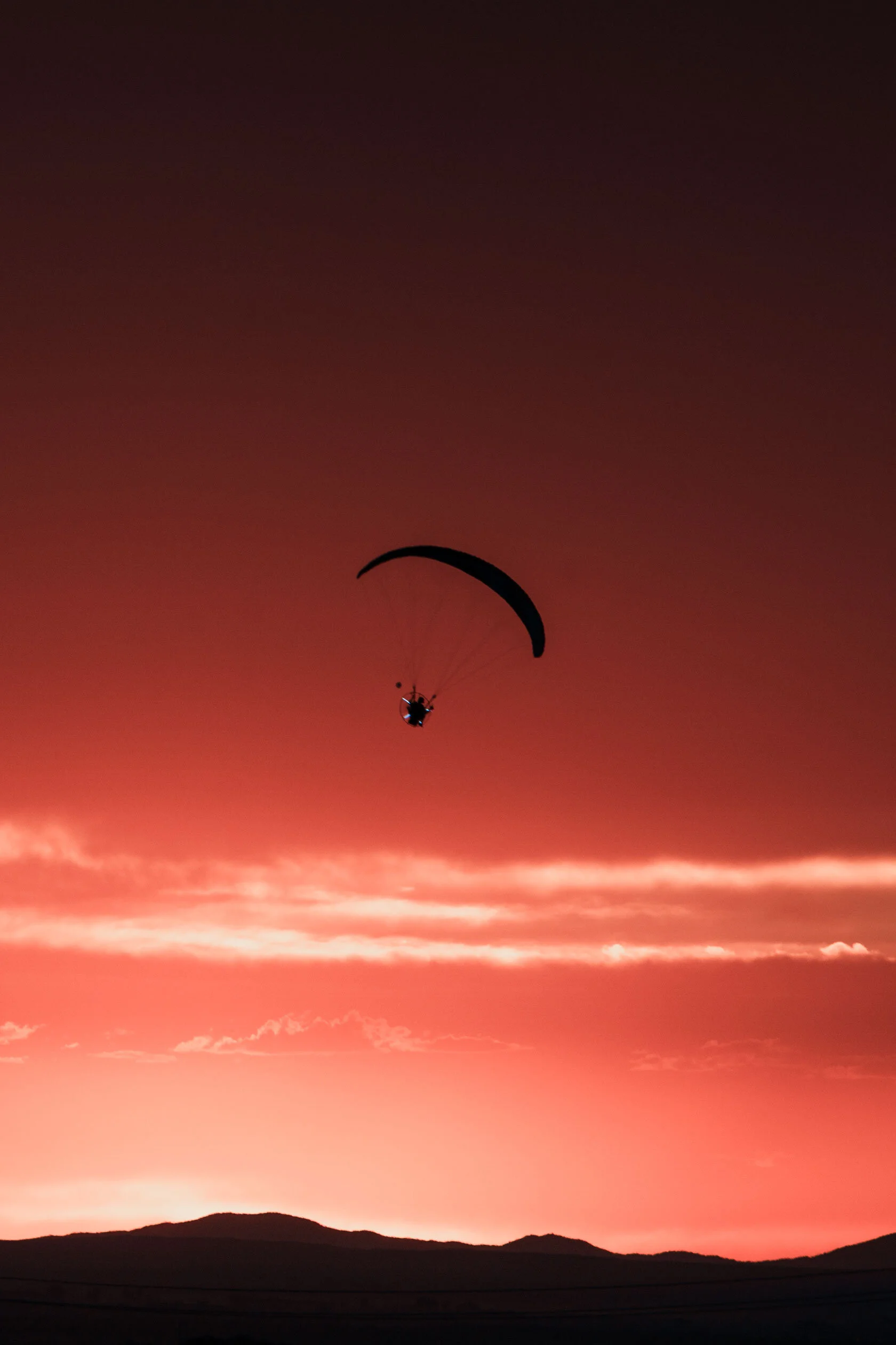Dark silhouette of guy wearing a motor with lights on it attached to paraglider flies over black, jagged mountains into colorful, dark blue to red orange gradient sunset. In Pocatello, Idaho.