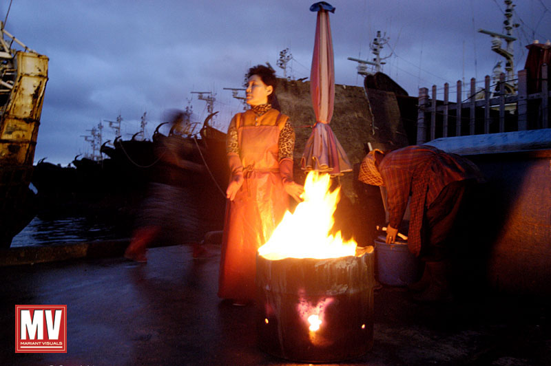 With the fishing fleet anchored in the background, women stay warm by a barrel fire before the sunrise opening of the fish market in Busan, South Korea.
