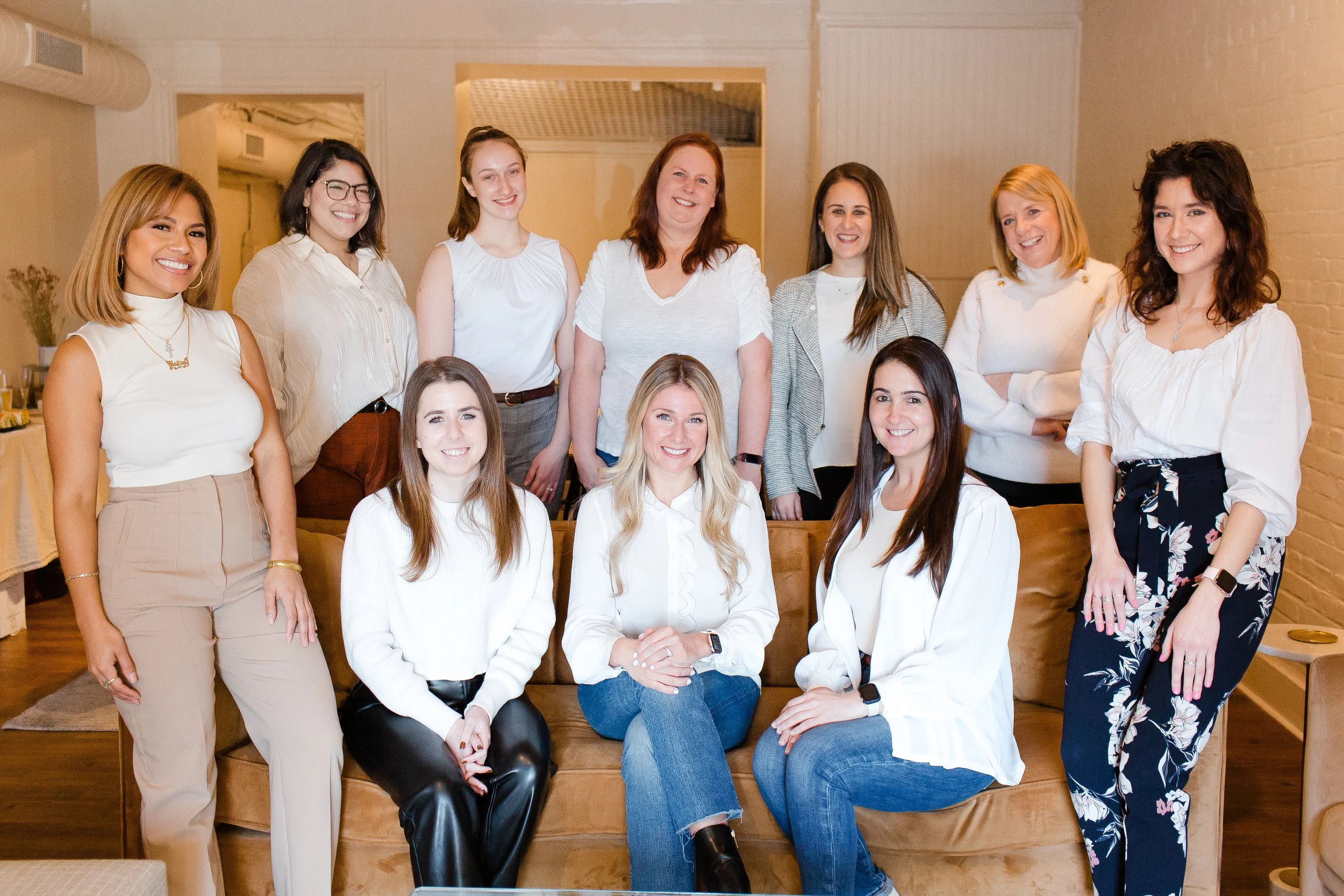 A group of twelve women posing together in a well-lit indoor setting, some seated on a sofa and others standing behind them, all smiling at the camera.