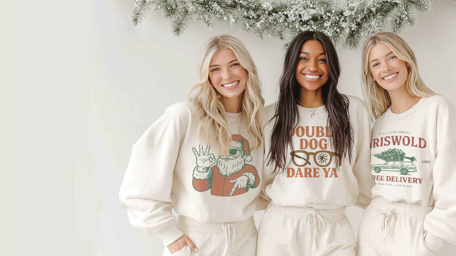 Three women smiling and standing close together, wearing casual holiday-themed sweatshirts, with a snowy pine branch overhead in the background.