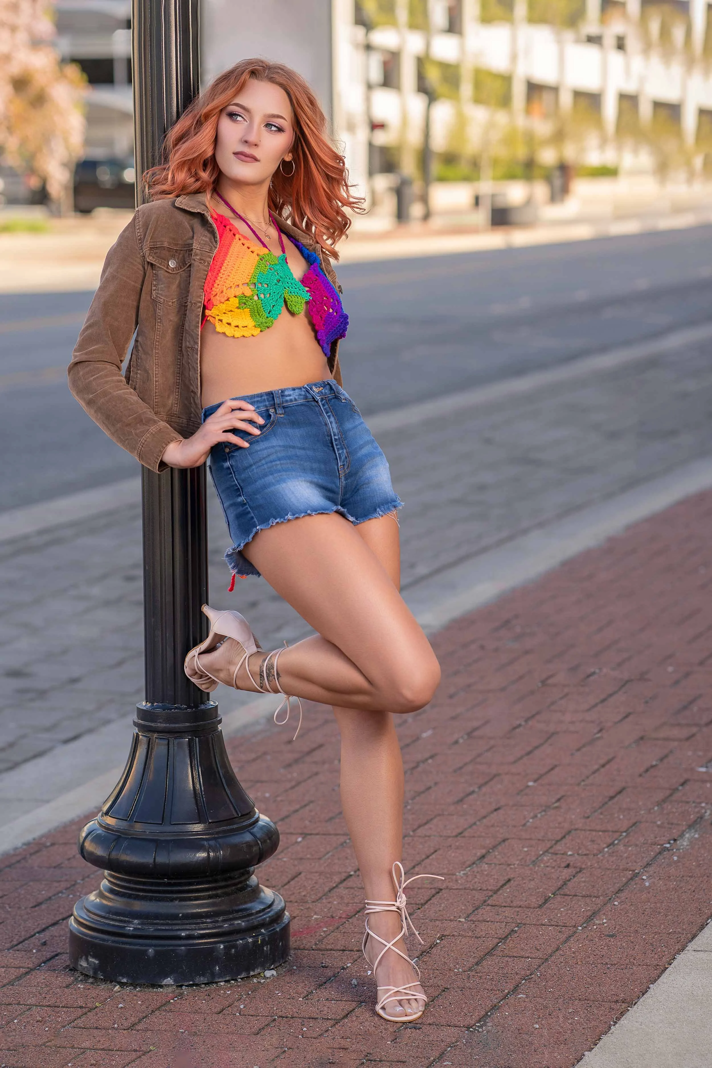 Lifestyle fashion shot of a woman in a rainbow crochet top and denim shorts leaning against a street lamp in Southern Michigan.
