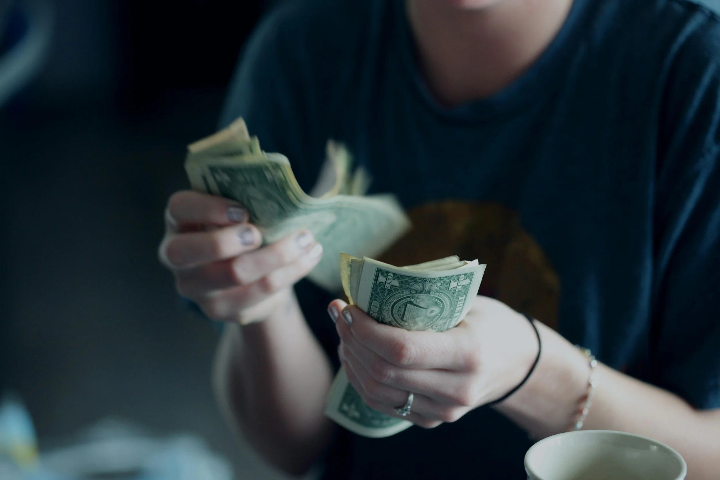Person counting and handling US dollar bills in a dimly lit environment, with a cup in the foreground.