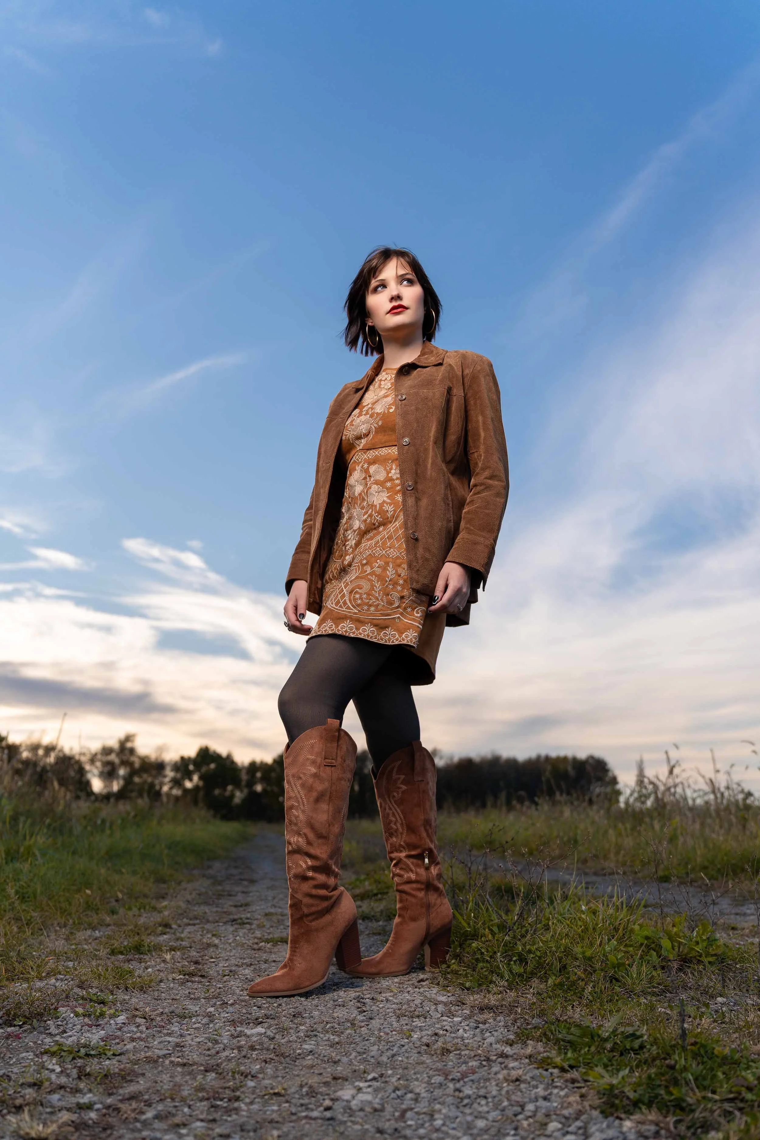 Full-length fashion portrait of a woman in tall brown boots and a suede jacket on a gravel path in Northern Indiana.