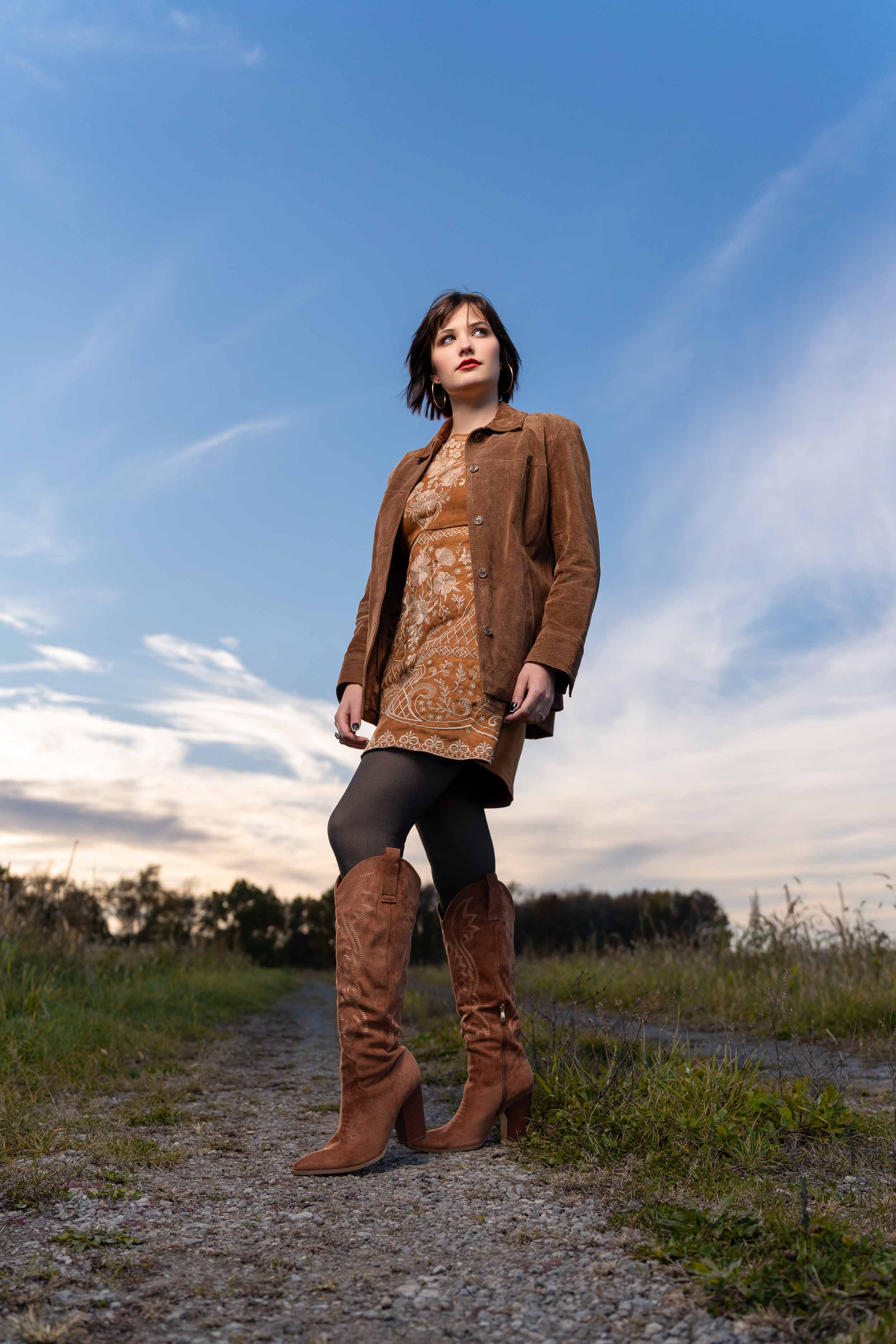 Outdoor lifestyle senior photography of a girl in a brown suede jacket and tall boots in a rural Southern Michigan field at sunset.