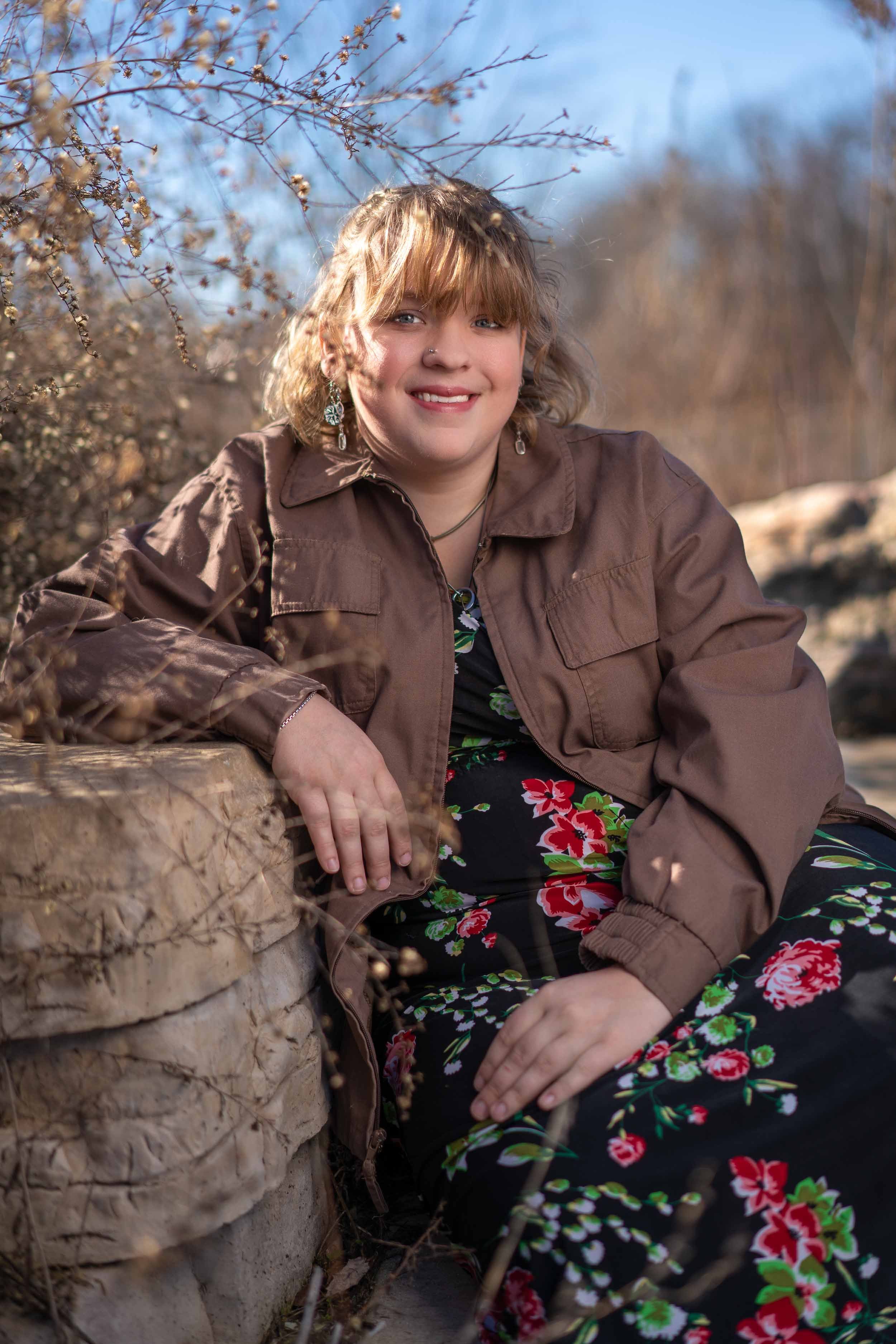 A smiling young woman in a floral dress and brown jacket sitting on a stone wall during an outdoor portrait session in Mishawaka.