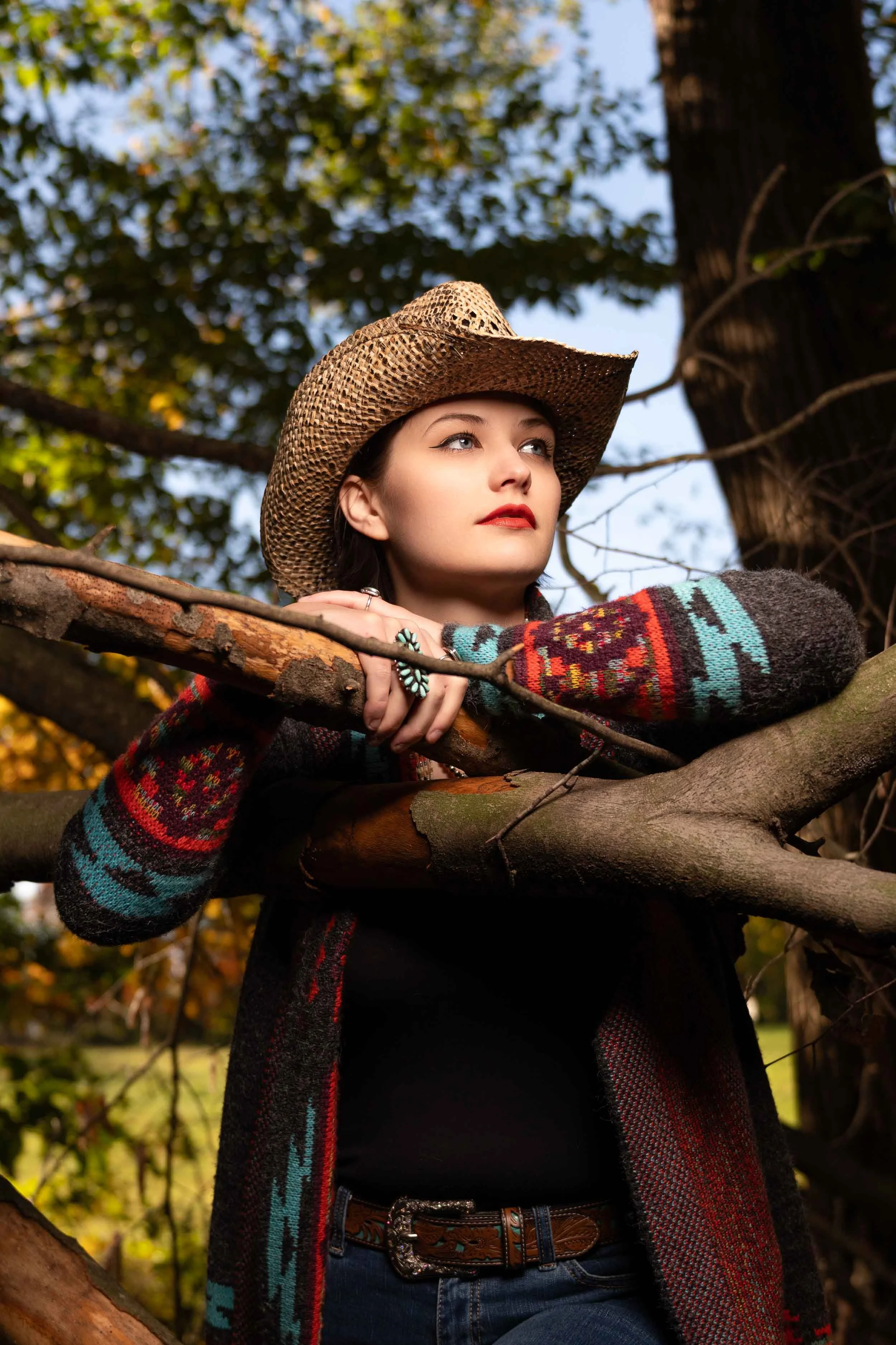 Western-inspired portrait of  young woman in a cowboy hat and patterned cardigan, leaning on a tree branch in a Northern Indiana park.