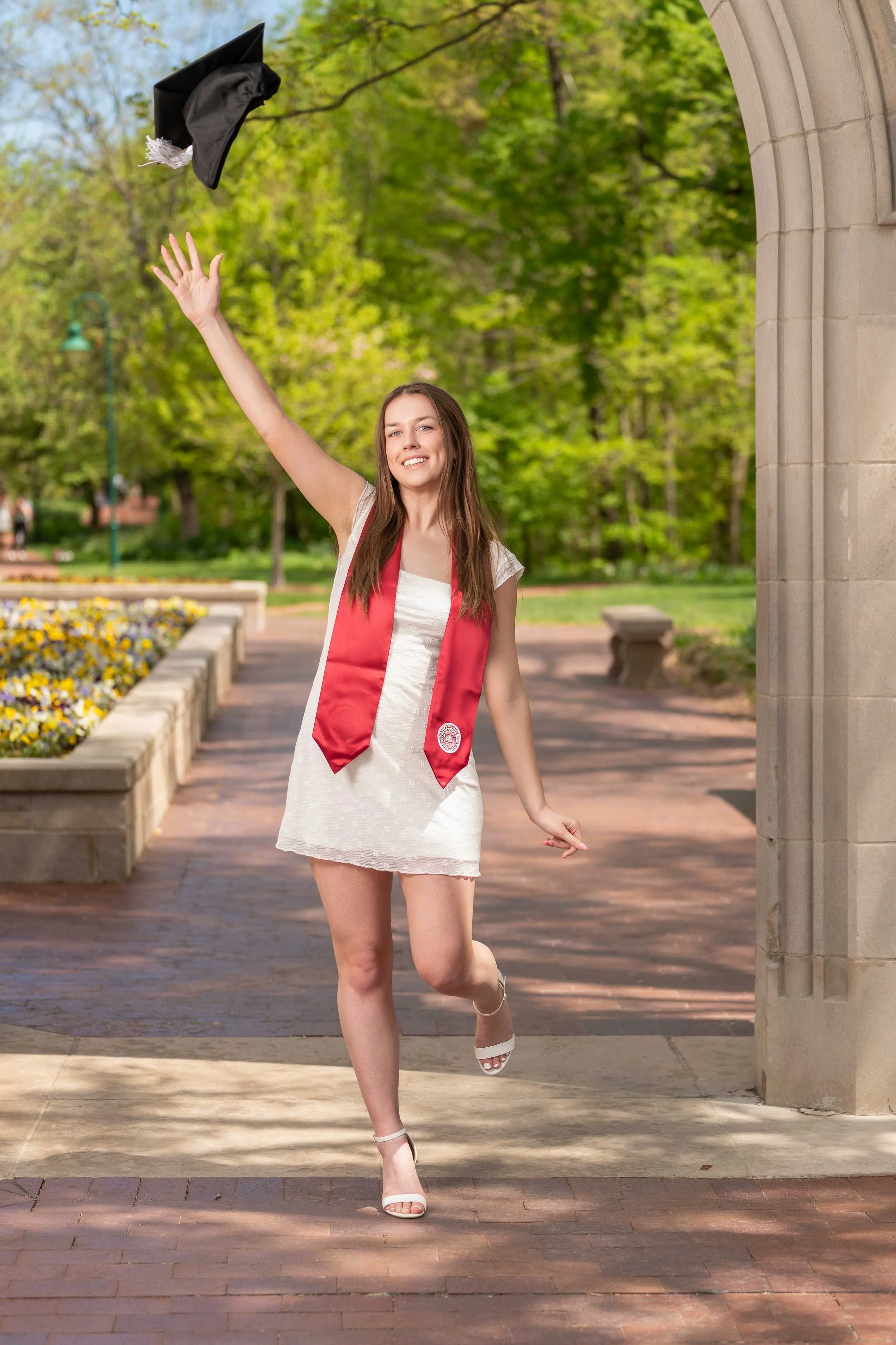 A college graduate in a white dress and red stole tossing her graduation cap in the air at a sunlit college campus in Indiana.