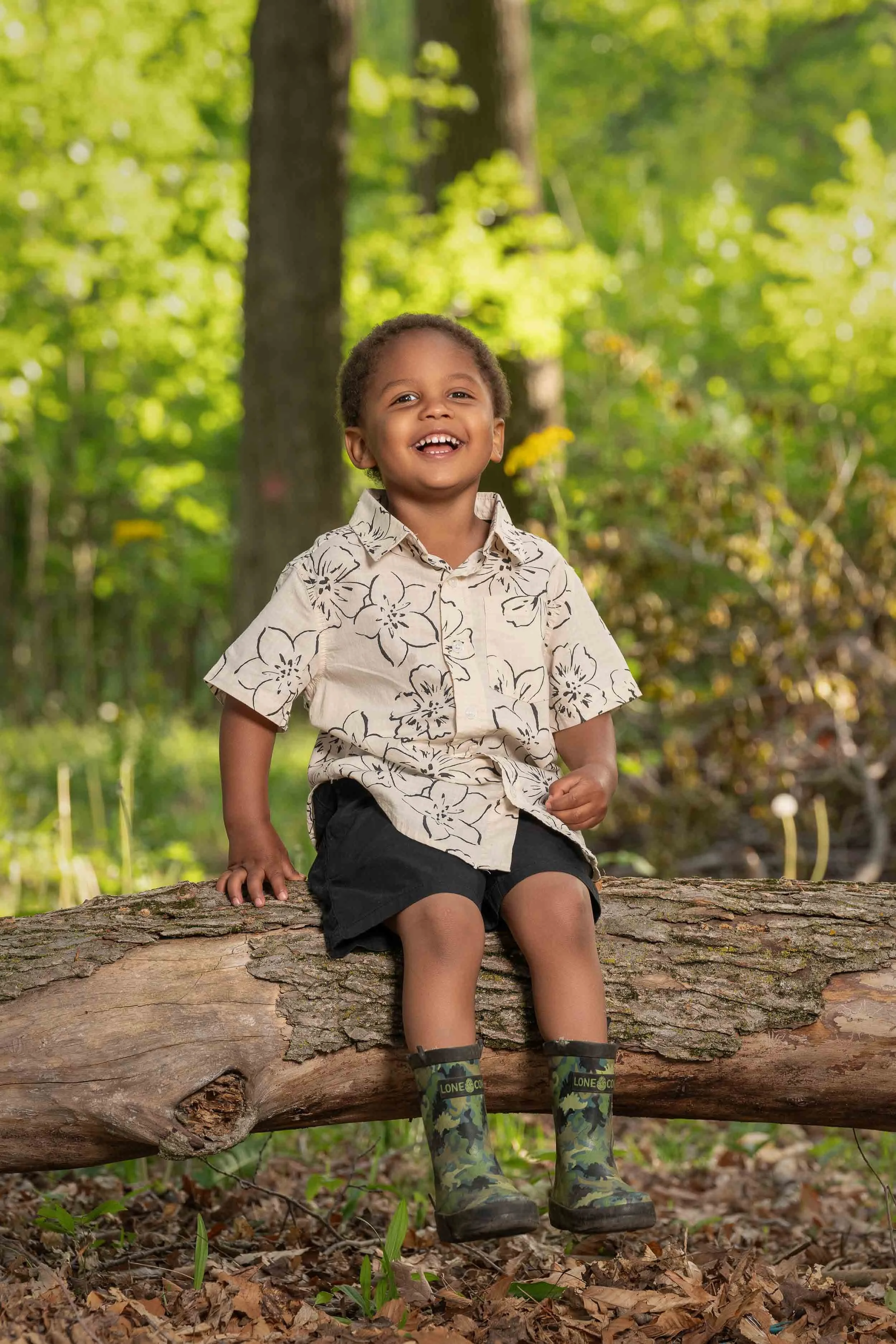 Young boy laughing while sitting on a fallen log in a wooded park, showcasing outdoor lifestyle child photography in Southern Michigan and Northern Indiana.