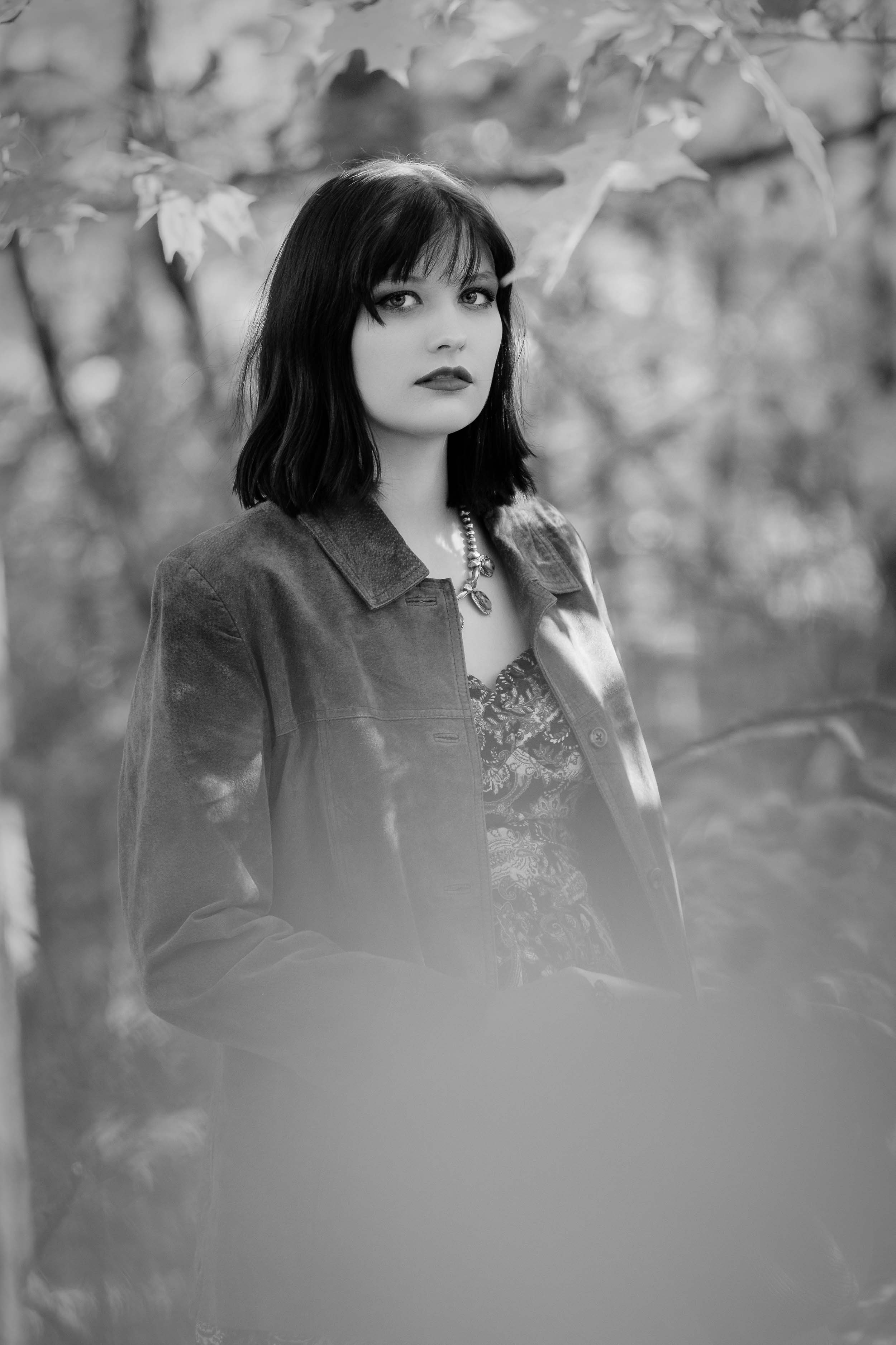 Moody fine art portraiture in black and white featuring a woman in a suede jacket among autumn trees in Northern Indiana.