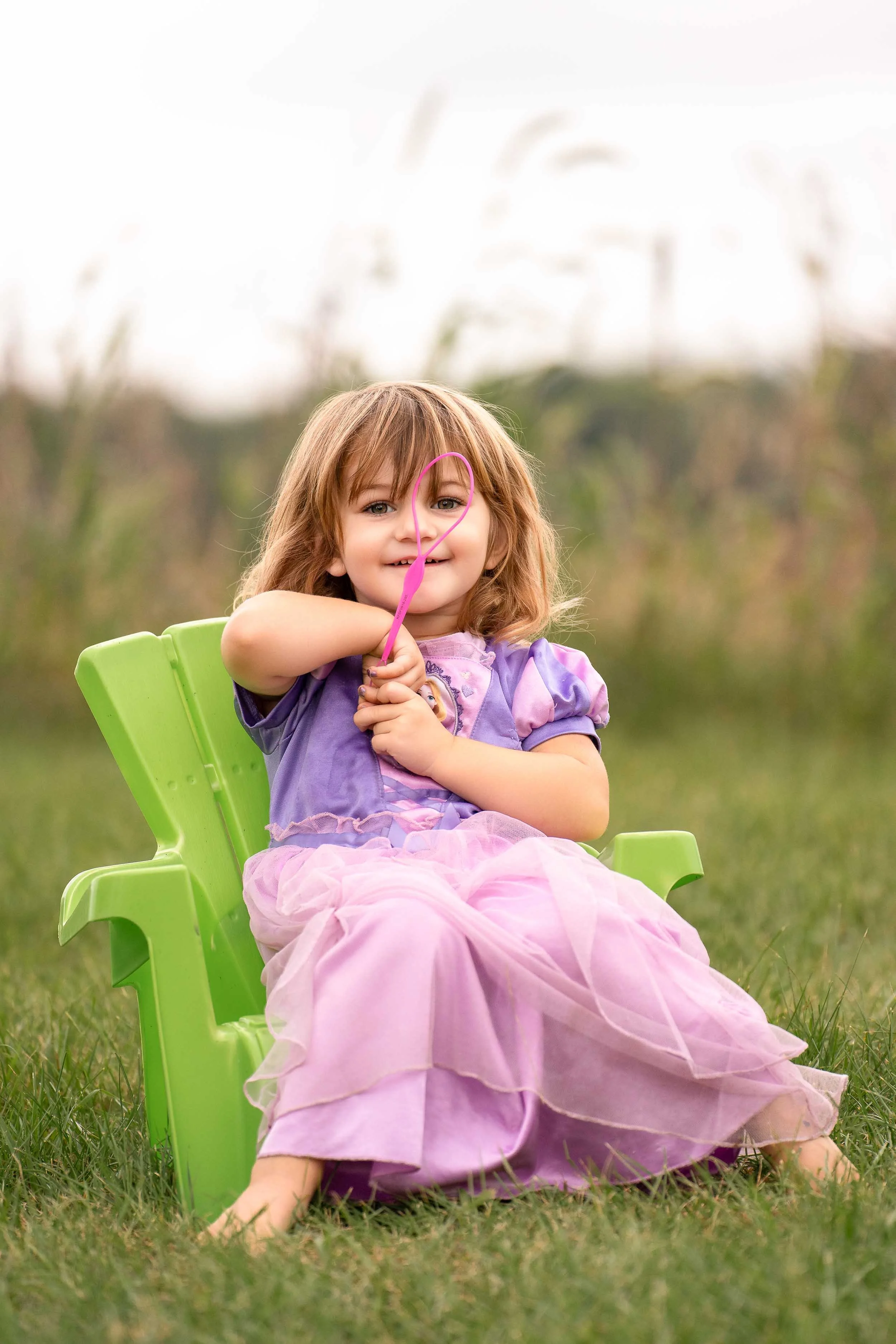 Toddler girl in a purple princess dress sitting in a green chair outdoors, providing child portrait photography services in Granger and the Michiana area.