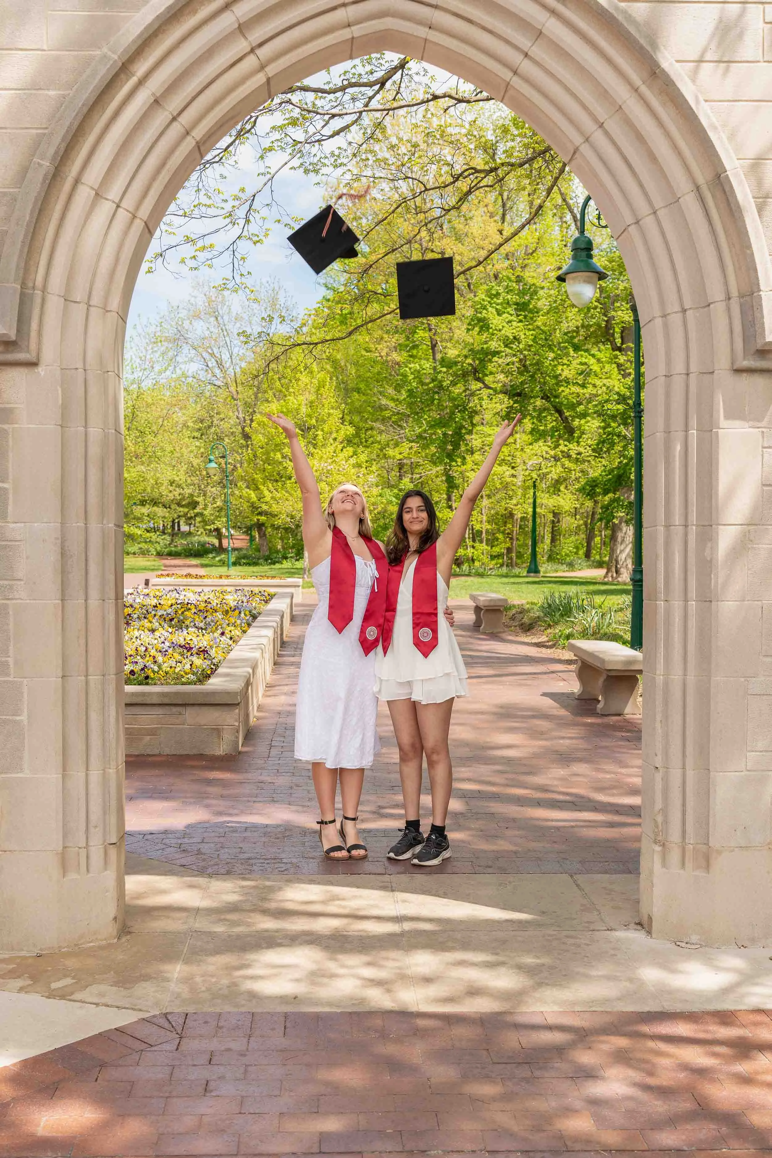 Two college seniors in white dresses and red graduation stoles throwing their caps in the air under a stone archway in South Bend, Indiana