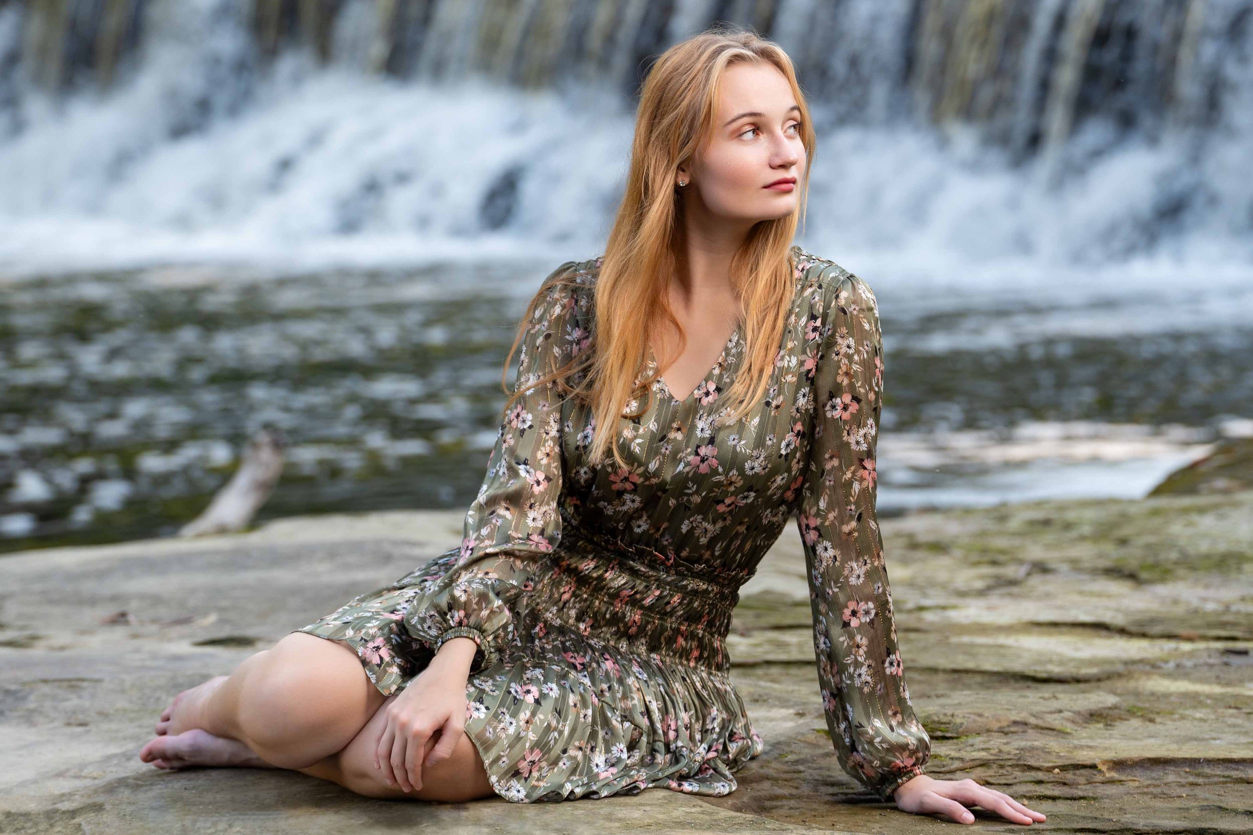 Professional outdoor portrait of a woman in a floral dress sitting by a waterfall in Northern Indiana.