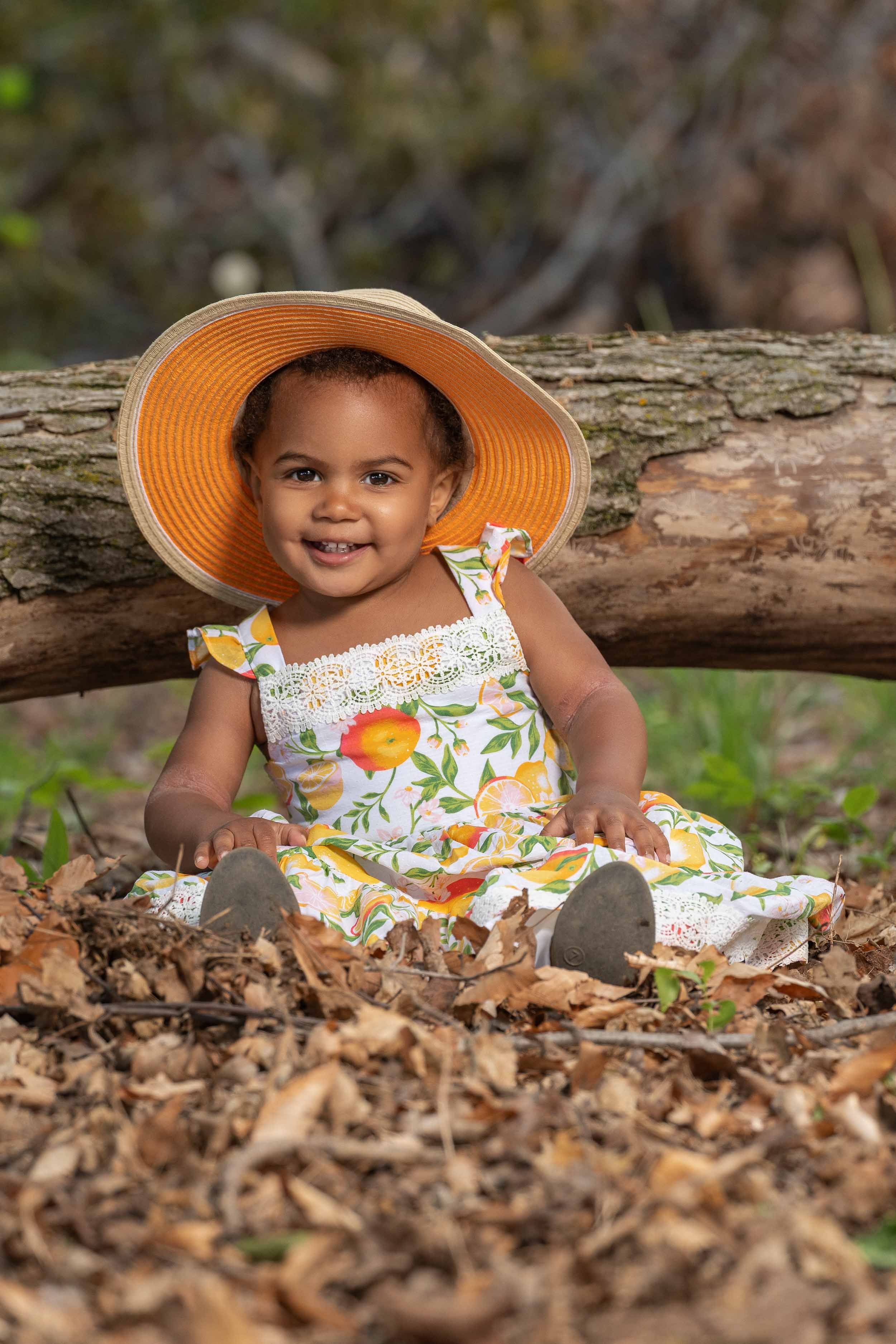 A young girl in a floral dress and a large sunhat sitting on the ground among fallen leaves with a log behind her, smiling at the camera.