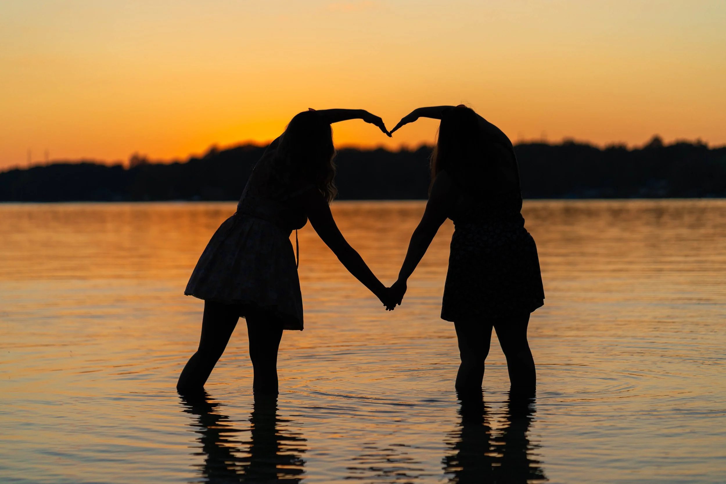 Silhouette of two best friends making a heart shape with their arms at sunset in a lake, captured by a Michiana senior photographer.