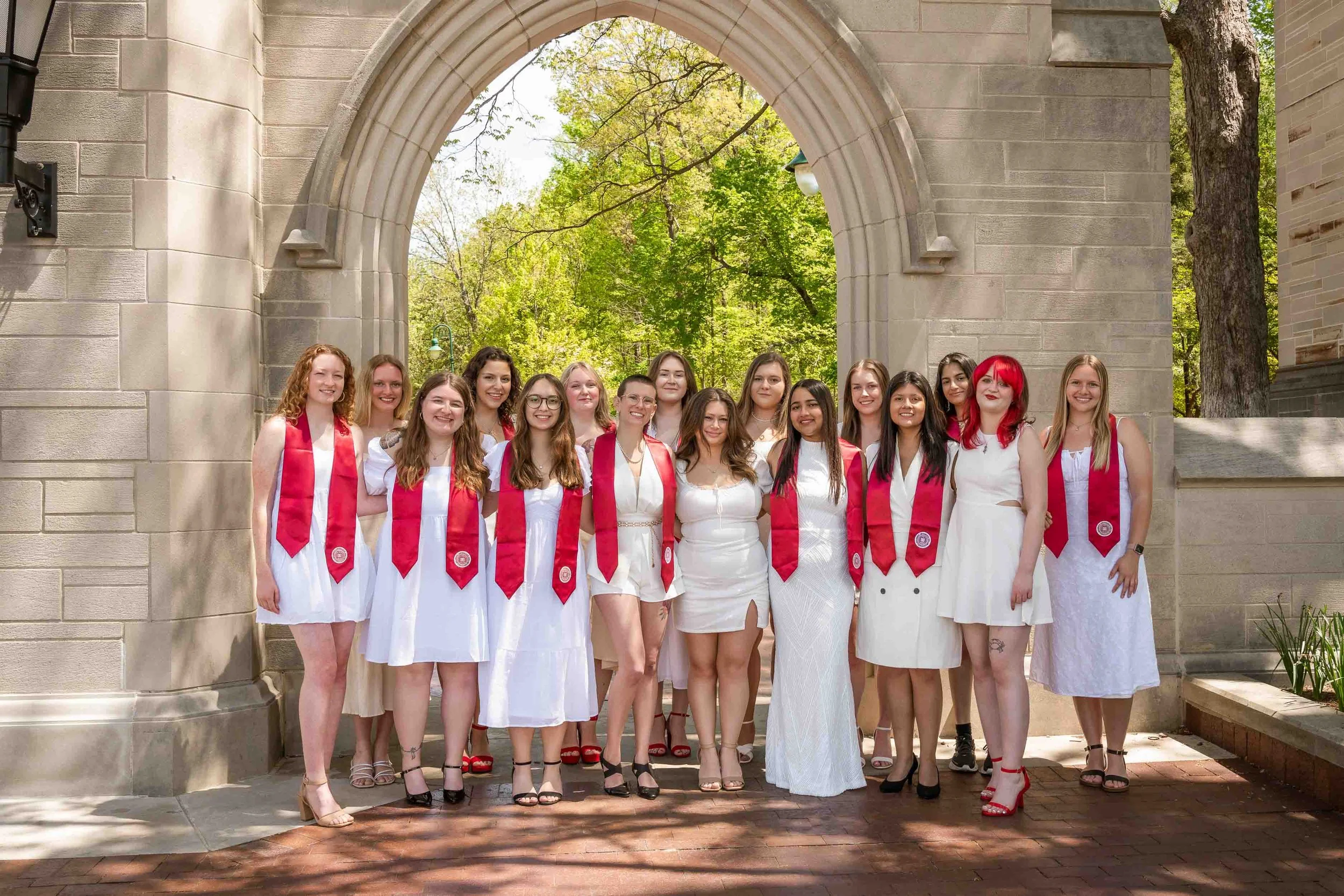 A large group of graduates in white dresses and red stoles posing under a stone archway; professional graduation photography by a South Bend senior photographer