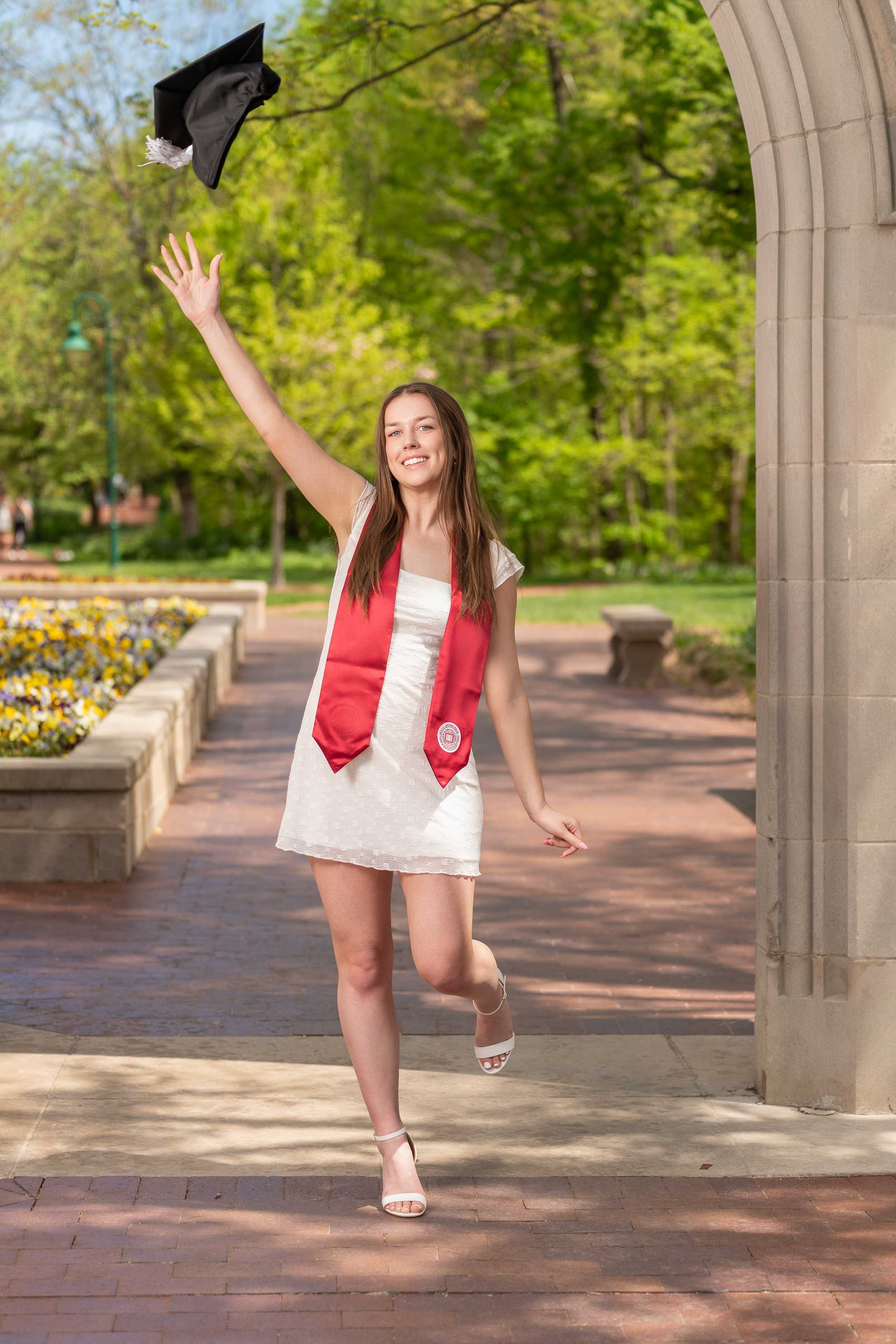 A  college graduate posing for a senior portrait in a white lace dress and red stole, tossing her graduation cap in a sunlit park setting in Northern Indiana