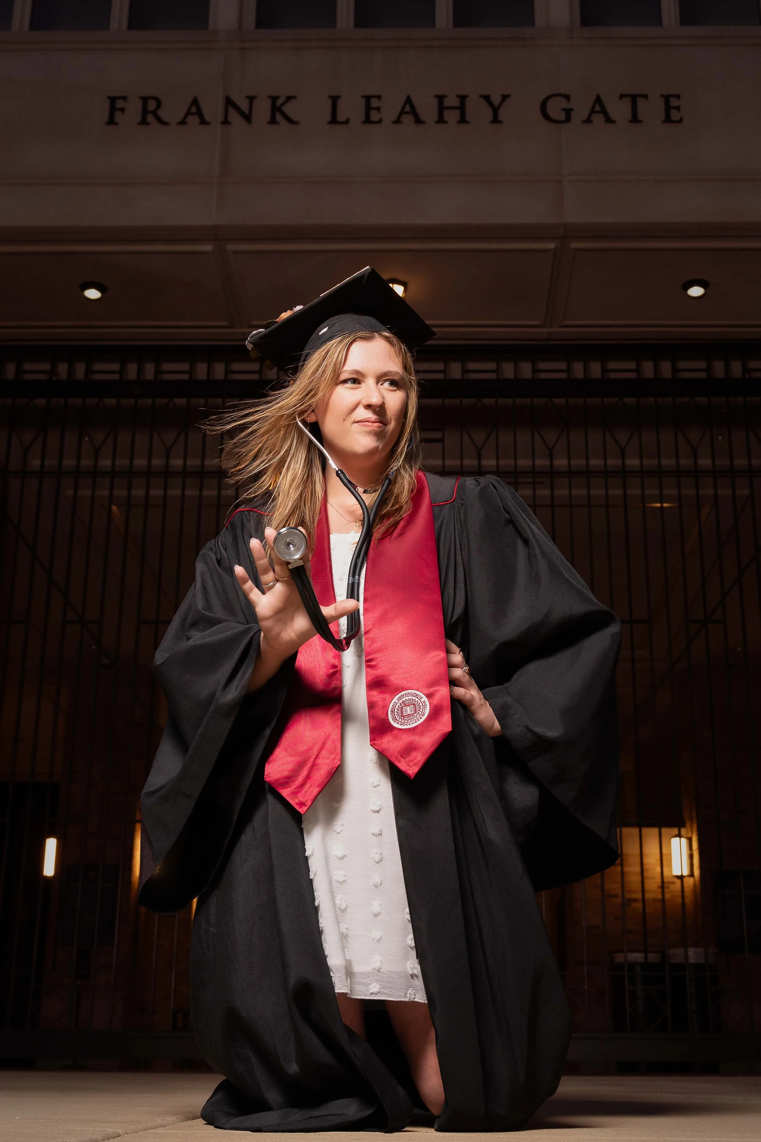 A nursing graduate in a black cap and gown holding a stethoscope in front of the Frank Leahy Gate at Notre Dame.