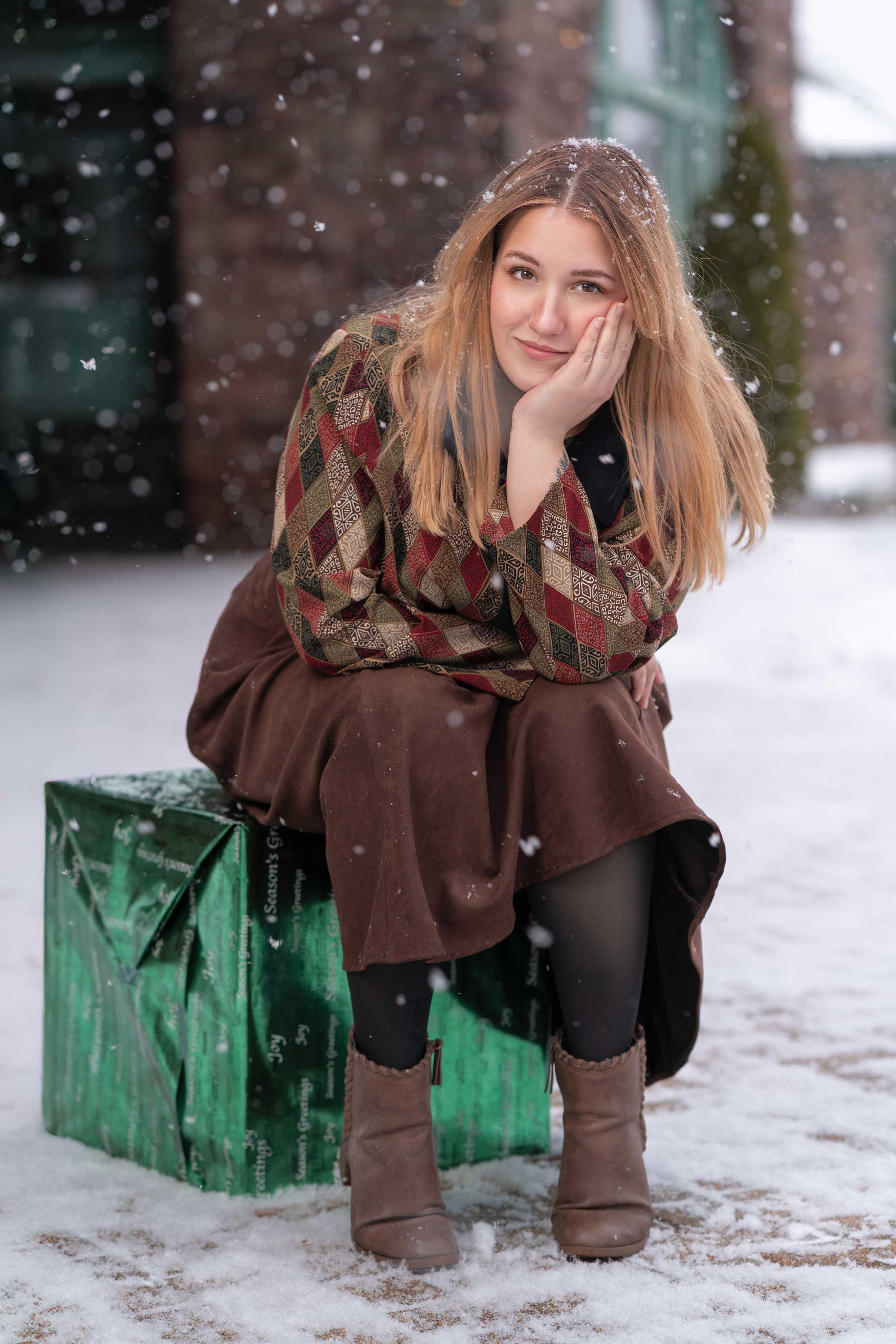 Winter lifestyle portrait of a woman sitting on a large green gift box during a snowfall in Southern Michigan, captured by James Russell Photography.