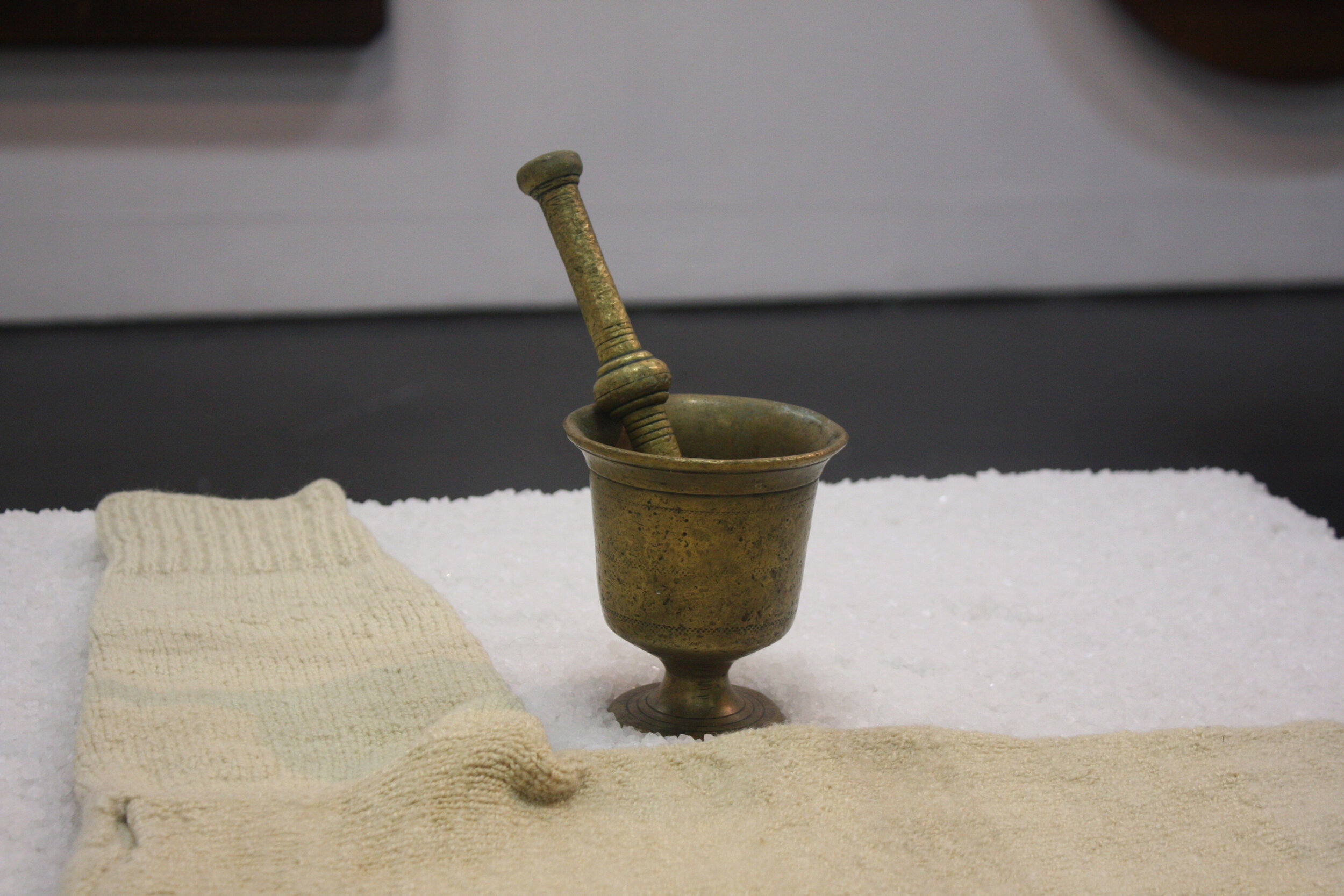    My sister’s table, my father’s garment, my mother’s mortar and pestle     (detail), 2013  Material: wooden table, granular salt, woolen handknitted garment, brass mortar and pestle. Presence: artist’s mother, father, sister, islands. 