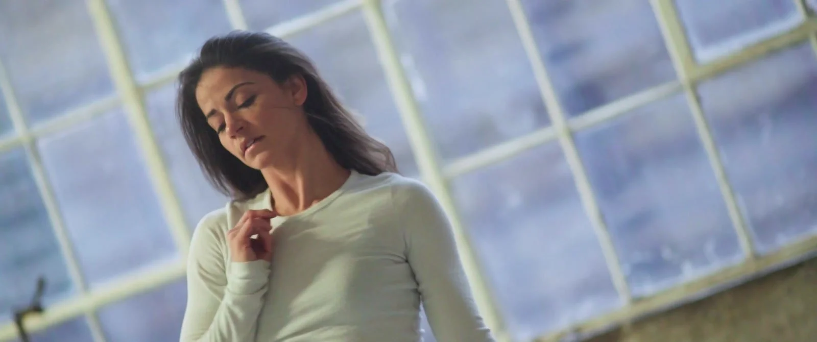 A dancer in a pale top moves in front of a large grid of sunlit windows.