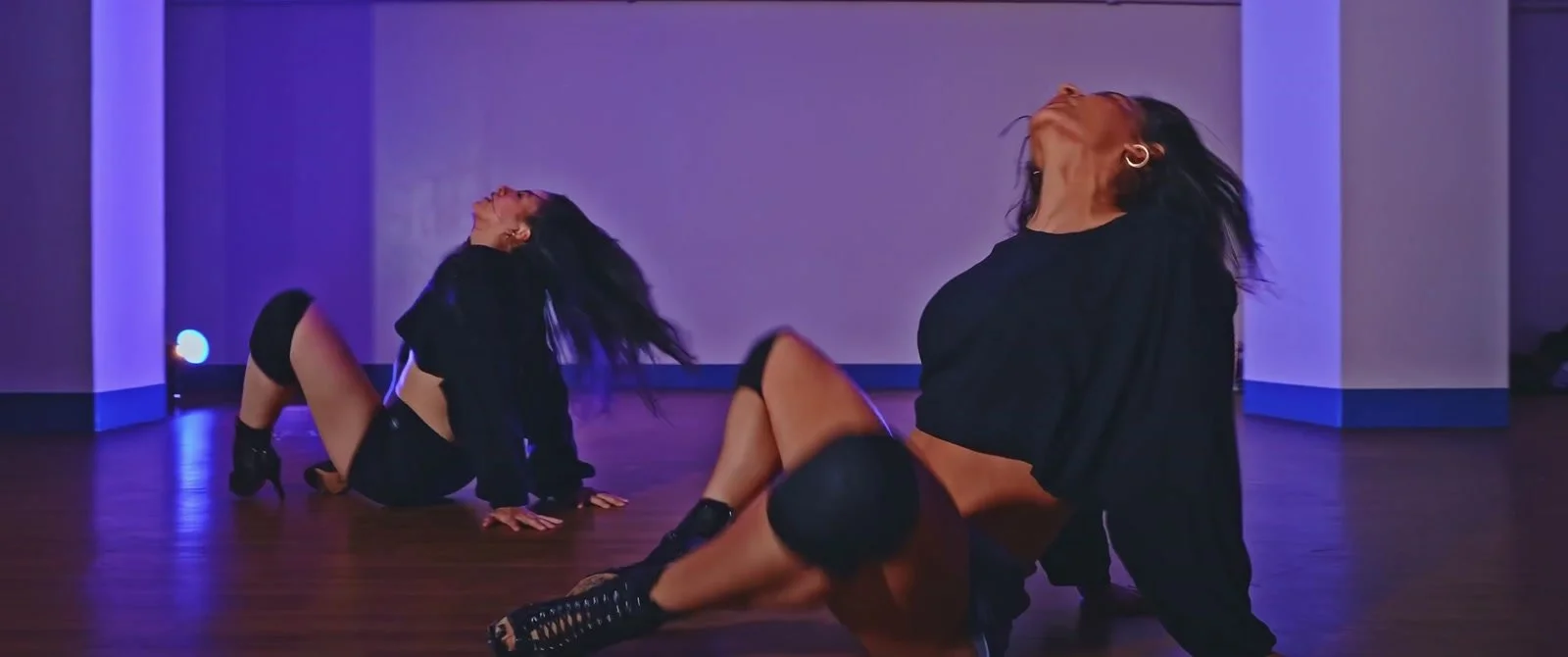 Two dancers lean back during a floor routine in a studio lit with violet light.