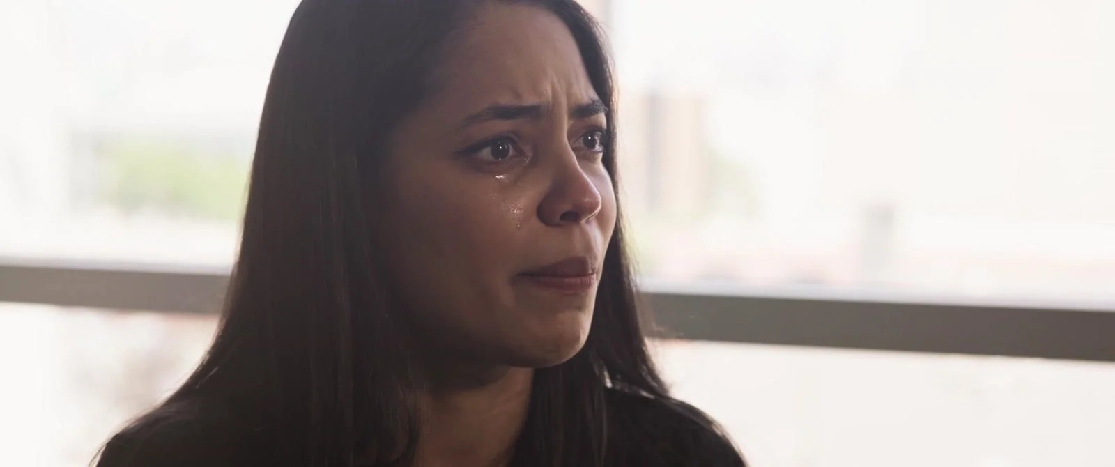A close-up of a woman with tears in her eyes beside a bright window.