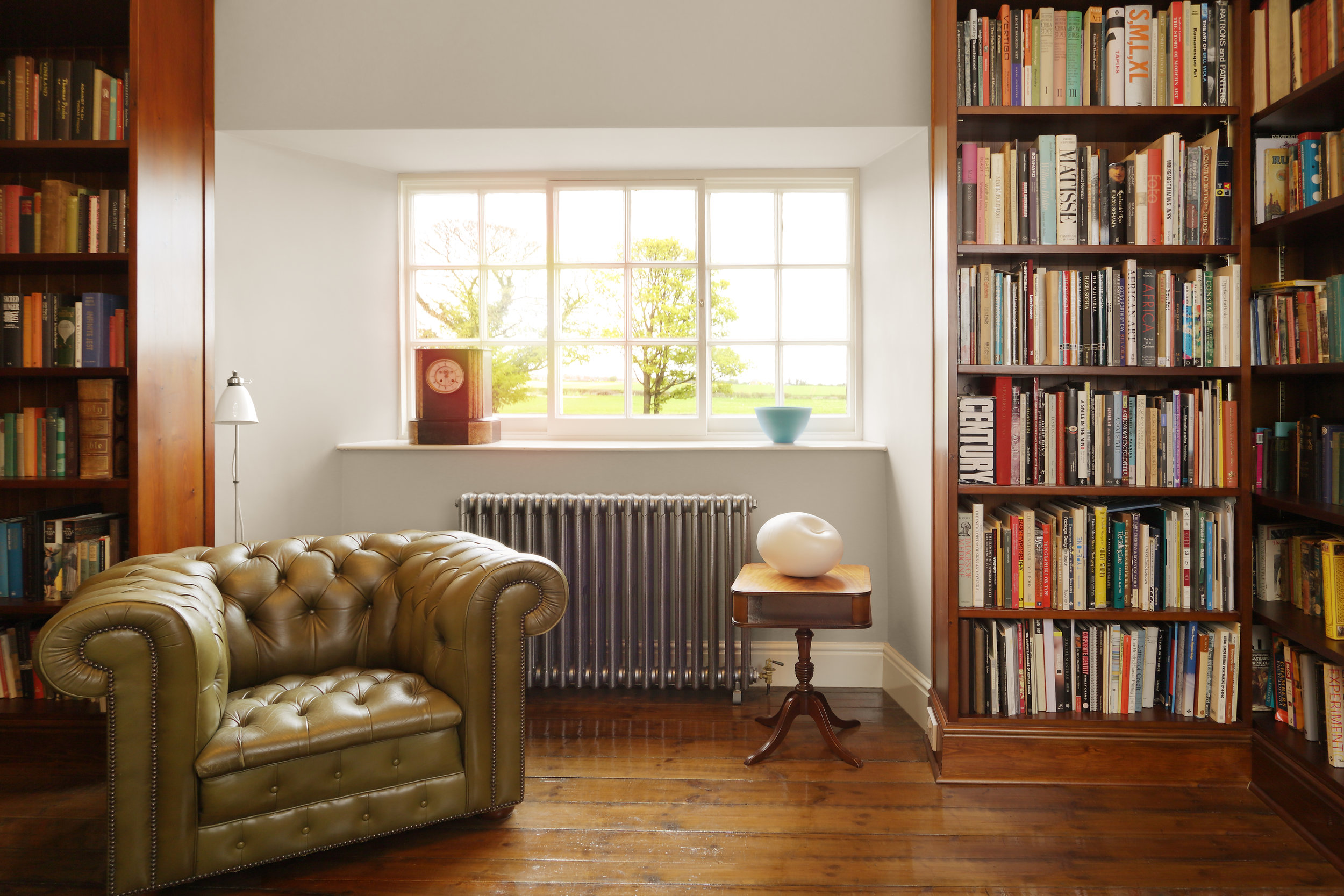  A metallic silver four column cast iron radiator in a relaxing library. A leather chesterfield armchair is surrounded by floor to ceiling bookshelves 