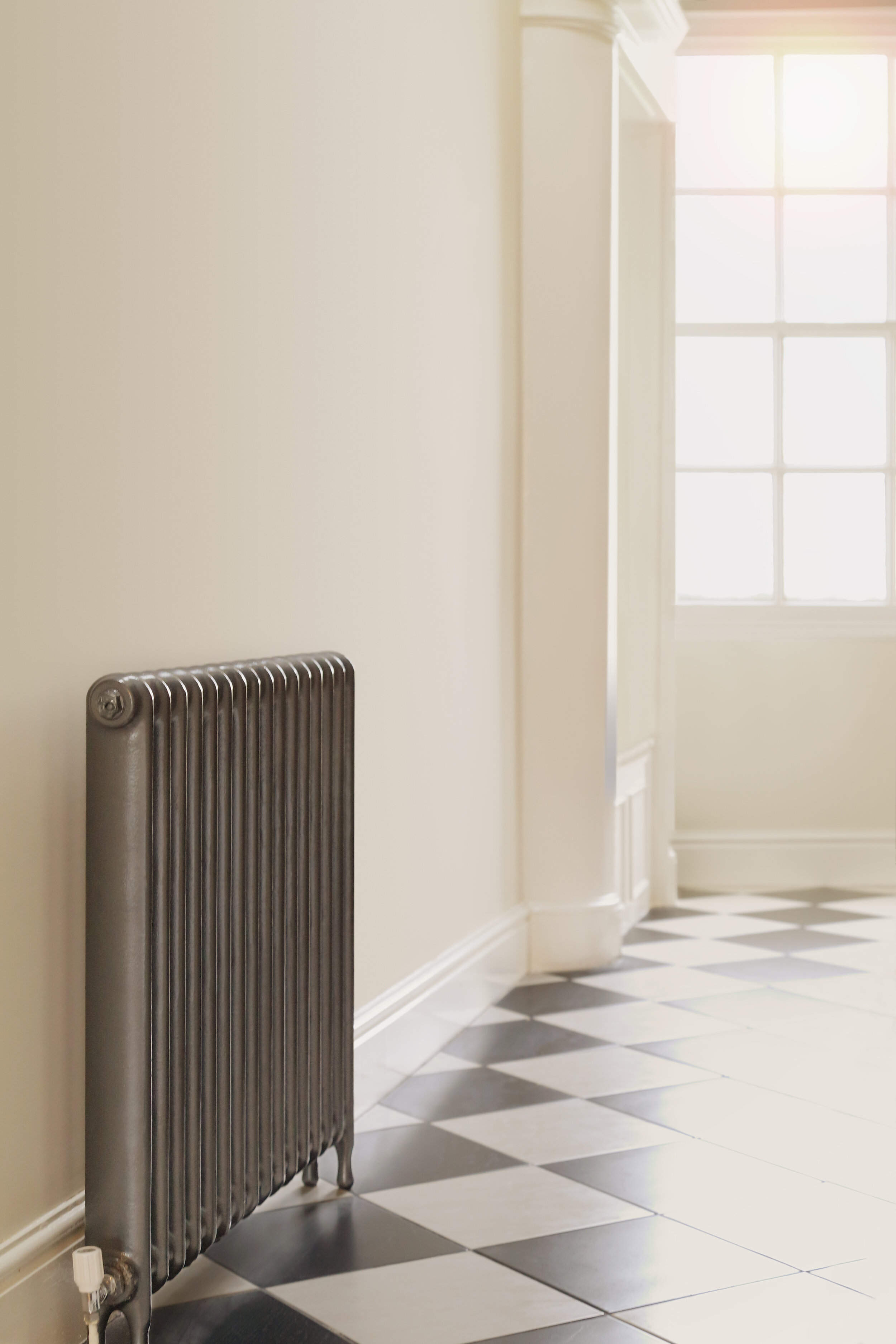  A narrow old school style cast iron radiator on a tiled hallway floor. A large window provides light 