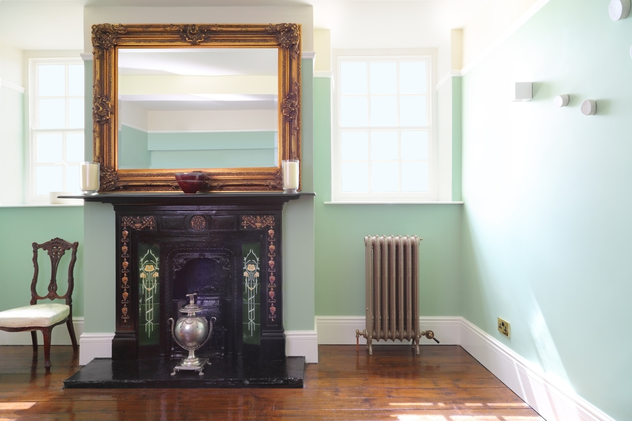  An ornate cast iron radiator on a wooden floor in a stately hall. The radiator is underneath a sash window and next to a very decorative fireplace with a silver urn in the centre. A large filigree mirror is hung above the fireplace 