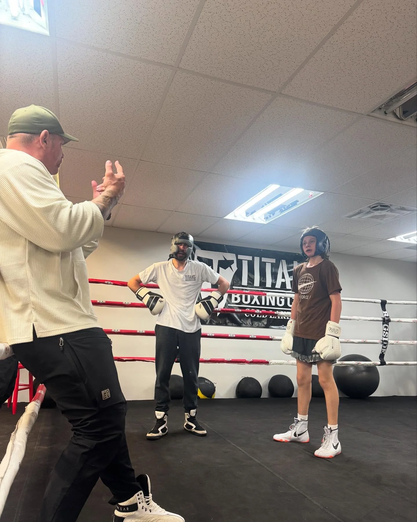 Boxing gym with four people: a coach on the left speaking and gesturing, two boxers in center and right in boxing gear, surrounded by a boxing ring with a 'Titan Boxing' banner on the wall.