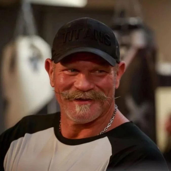 Smiling man wearing a black cap with unreadable text, a chain necklace, and a black and white shirt, in a workshop setting.