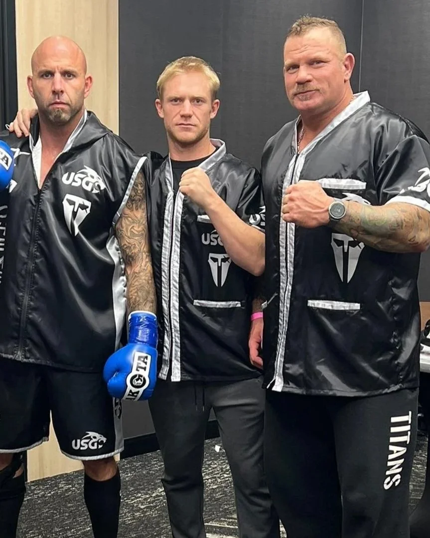 Three men in black boxing jackets, with the middle man making a fist, posing in a room with black and beige walls.