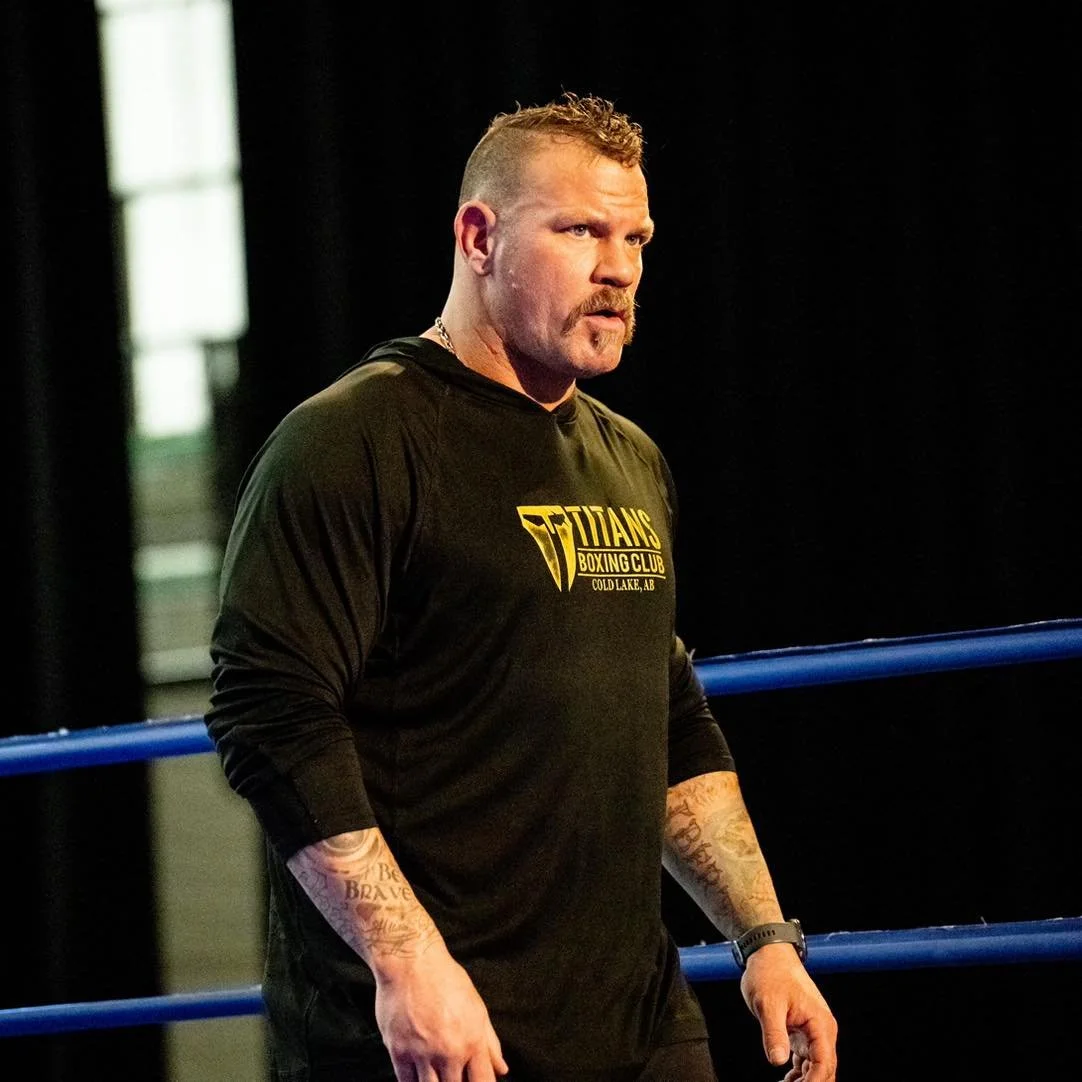 A serious man with short hair and tattoos on both forearms, wearing a black long-sleeve sweatshirt with yellow text, standing inside a boxing ring.