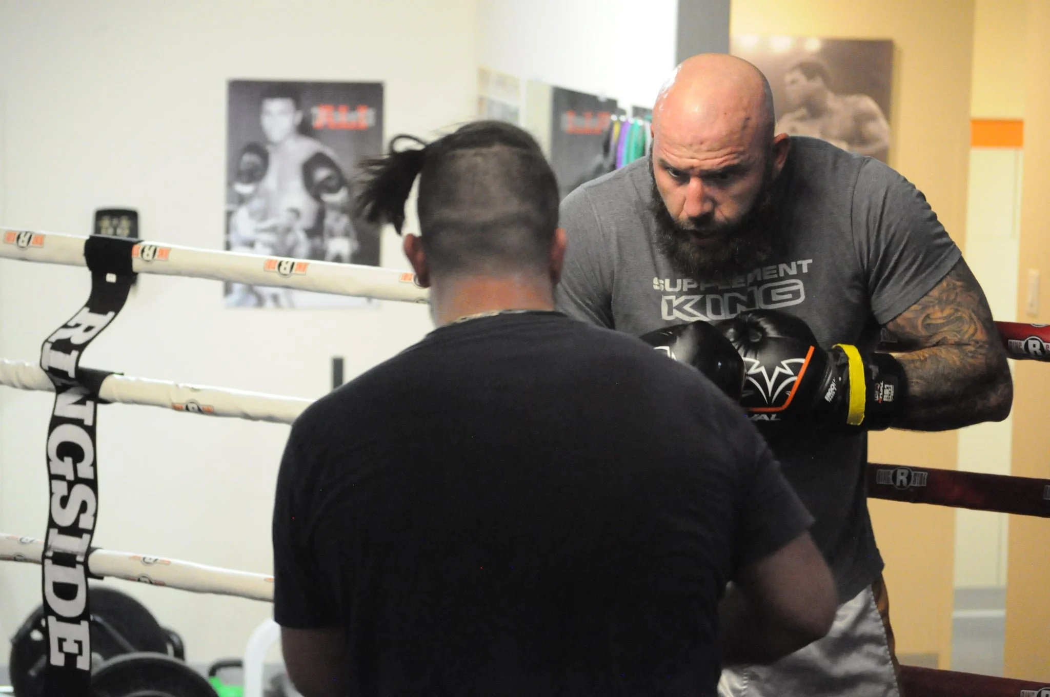 Adult male boxer with a tattoo on his arm practicing boxing with his trainer in a gym, standing inside a boxing ring with posters on the wall in the background.