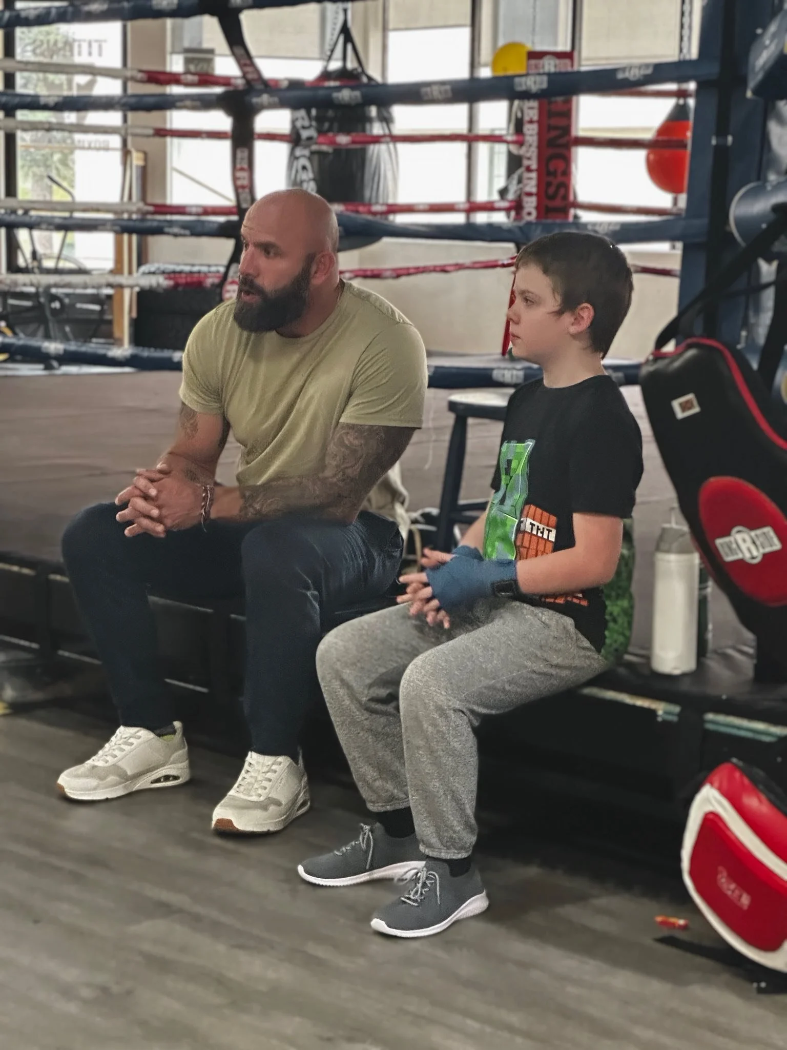 A man and a boy sitting on a bench inside a boxing gym near a boxing ring.