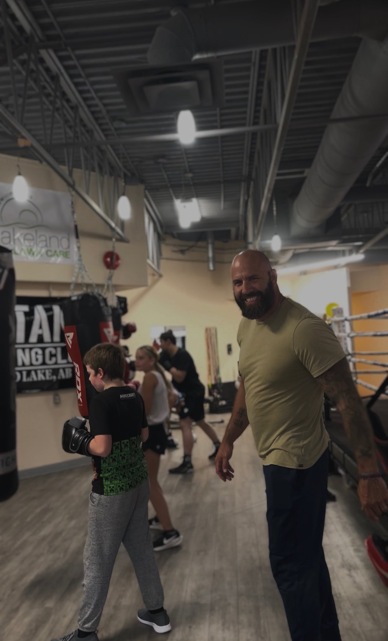 A man with a beard smiling in a gym, surrounded by children practicing boxing with punching bags and workout equipment.