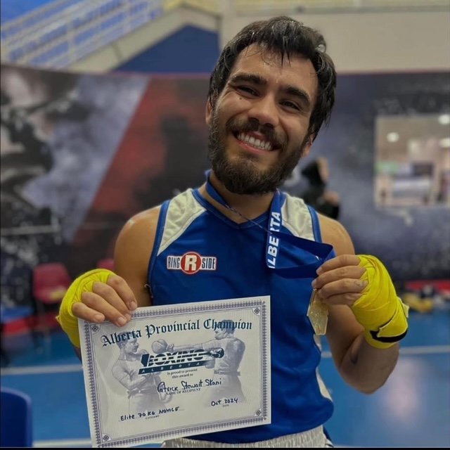 A smiling man in a blue sleeveless athletic shirt holding a certificate and a medal, wearing yellow gloves, in an indoor sports facility.