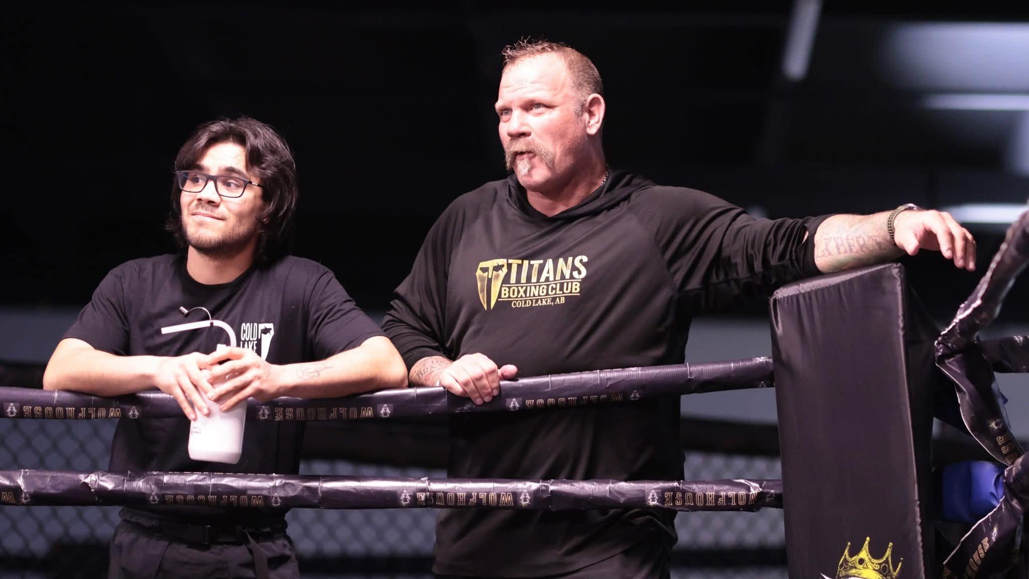 Two men standing inside a boxing ring, one wearing a Titans Boxing Club shirt and the other wearing a Cold Lake shirt, leaning on the ring ropes.