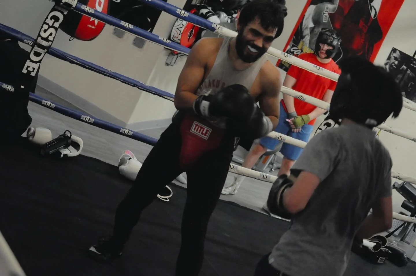 A man with a beard and dark hair wearing a sleeveless shirt, black gloves, and black pants is smiling and holding boxing pads inside a boxing gym. Behind him, a young person with glasses, dark hair, and a gray shirt is holding gloves, and another per