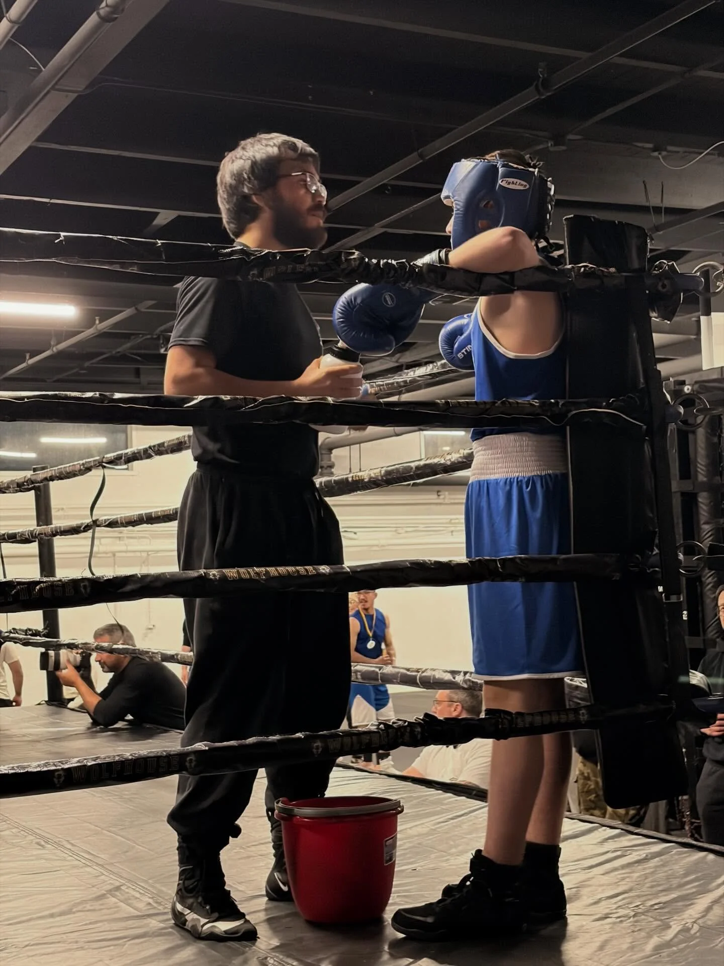 A boxing trainer coaching a young boxer in a boxing ring with ropes, in an indoor gym. The trainer is holding water bottles, and the boxer is wearing protective headgear and boxing gloves.