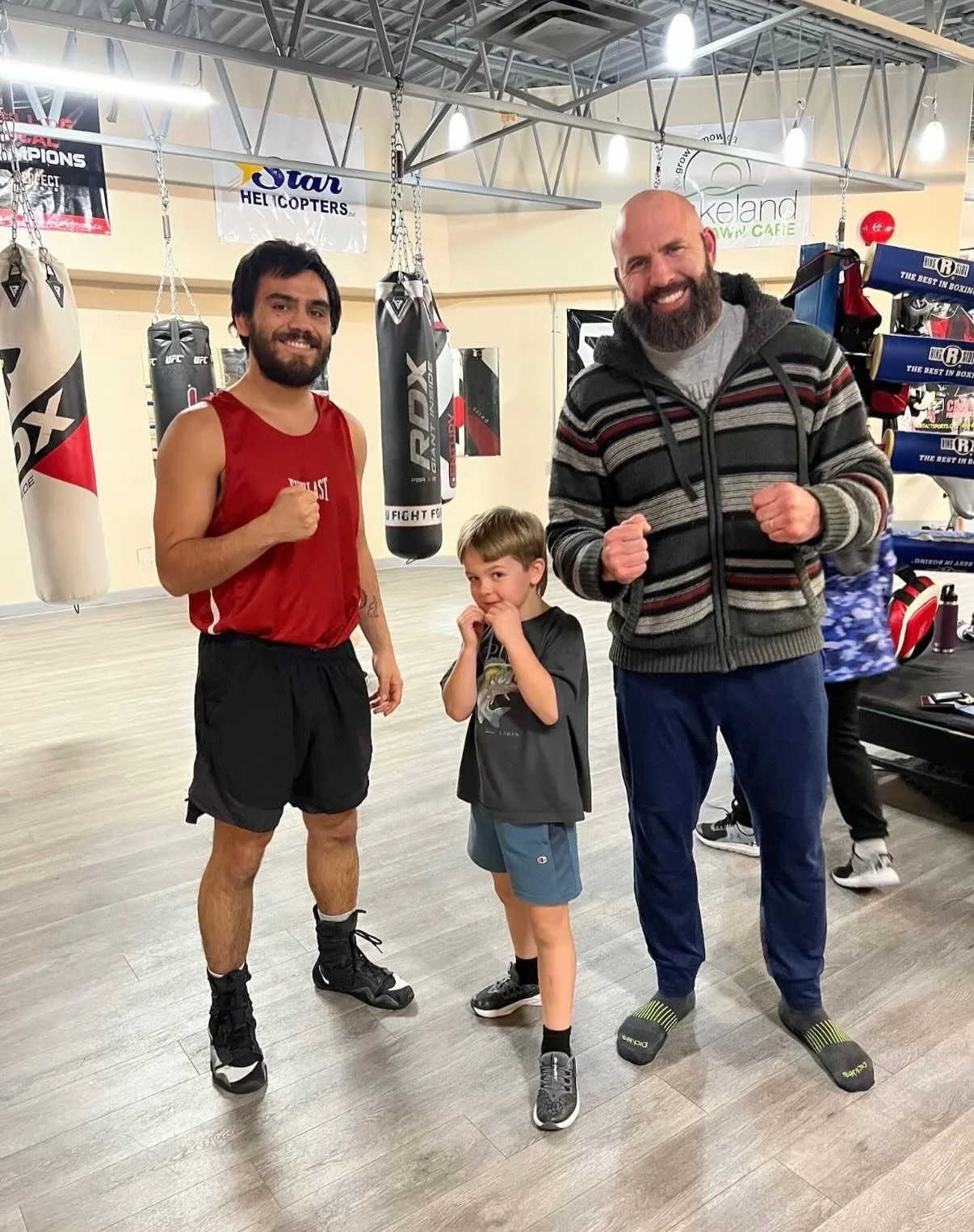 Three people, a boxer, a young boy, and a man, standing in a boxing gym with punching bags hanging behind them, all smiling and showing a fighting stance.