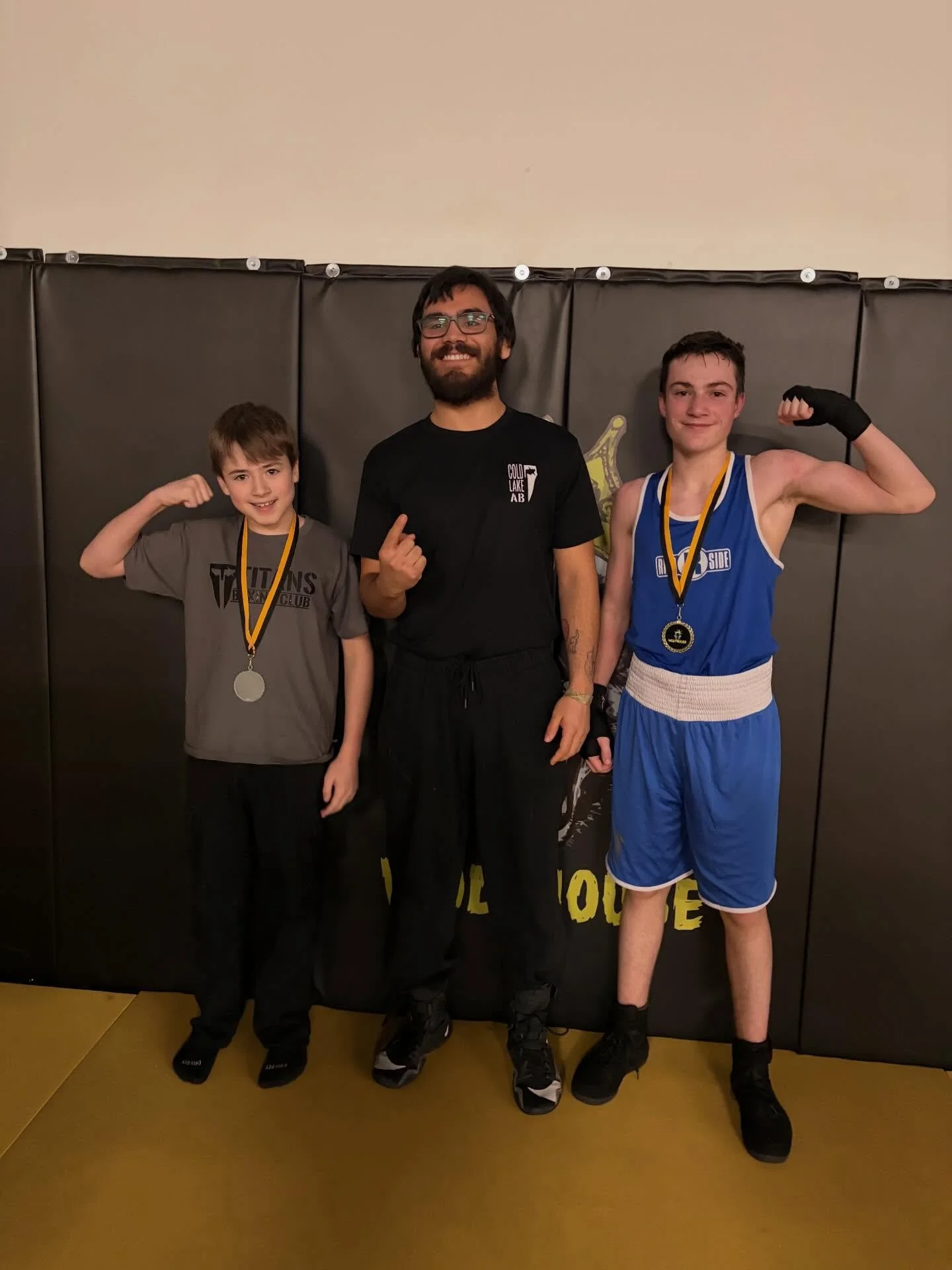 Two young boys and a man posing with medals, flexing their arms, in front of a black wall padding at a sports event.