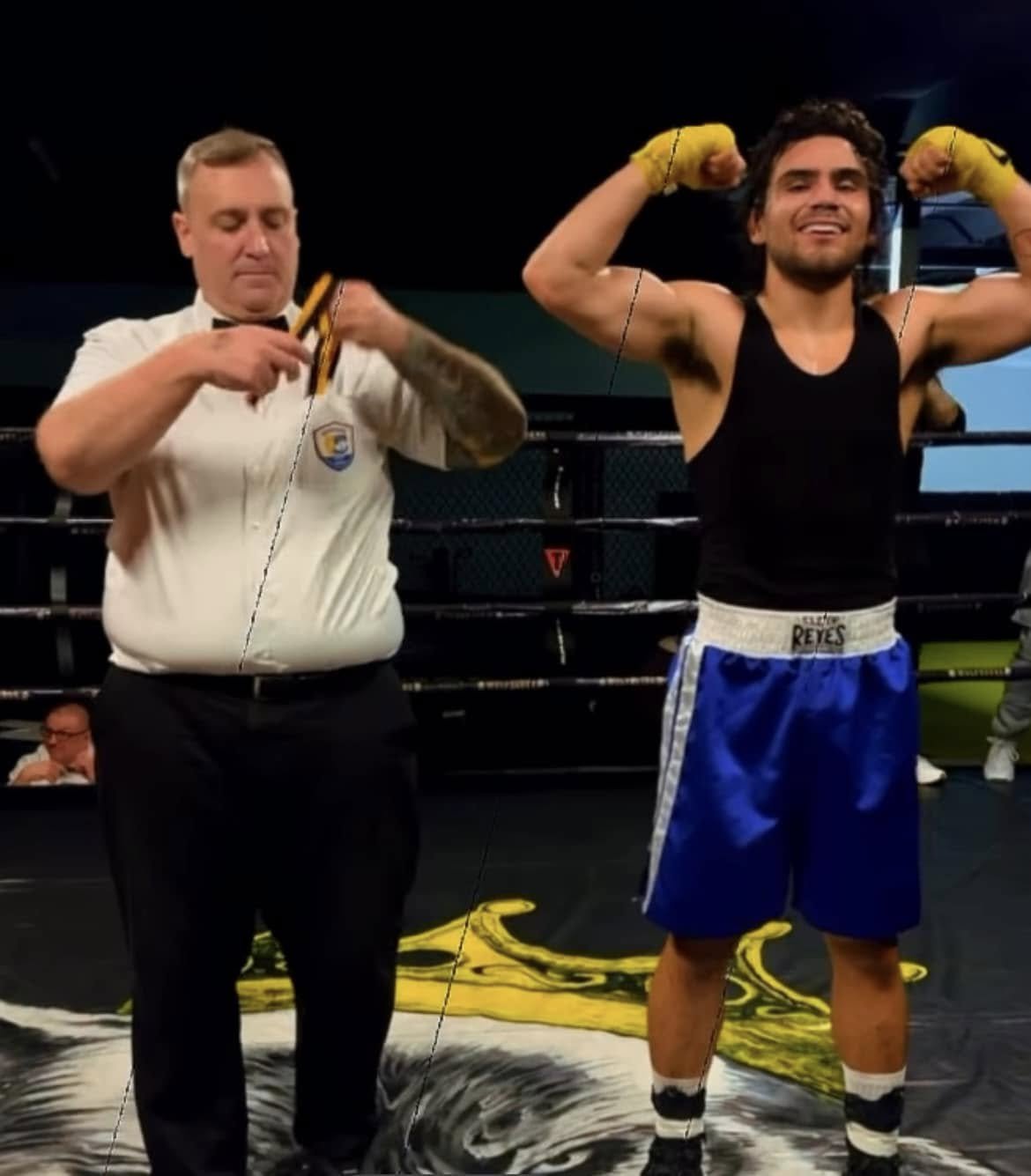A boxer with yellow gloves flexes his biceps in a boxing ring, while a referee dressed in a white shirt assists with his gloves.