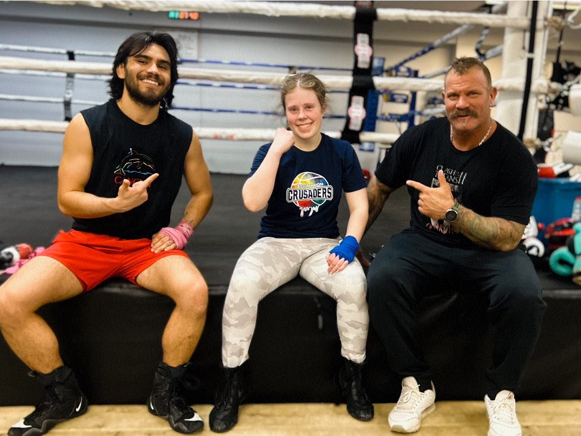 Three people sitting on a boxing ring floor smiling at the camera, with boxing gloves and equipment in the background.