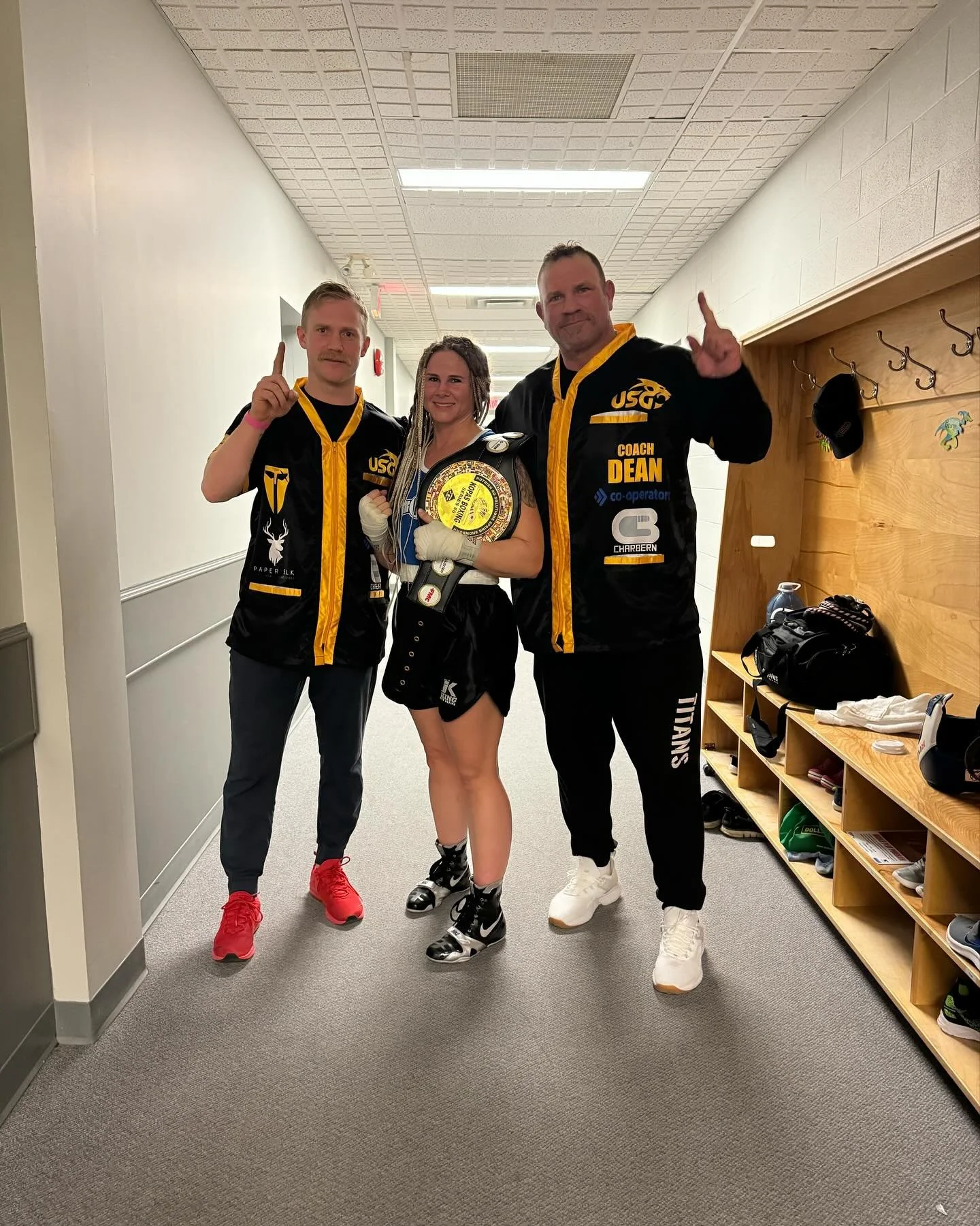 A victorious female boxer holding a championship belt, flanked by two male trainers or coaches, all in athletic attire with team jackets, posing in a hallway with wooden lockers and bags in the background.