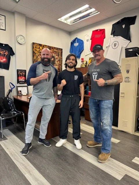 Three men standing together in an office space, smiling and making fist gestures. Behind them are shelves with sports shirts and memorabilia, a locker, and a wall decorated with artwork and a clock.