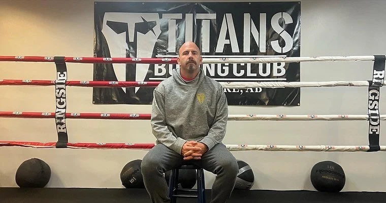Man sitting on a stool in front of a boxing ring with a Titans Boxing Club banner in the background.